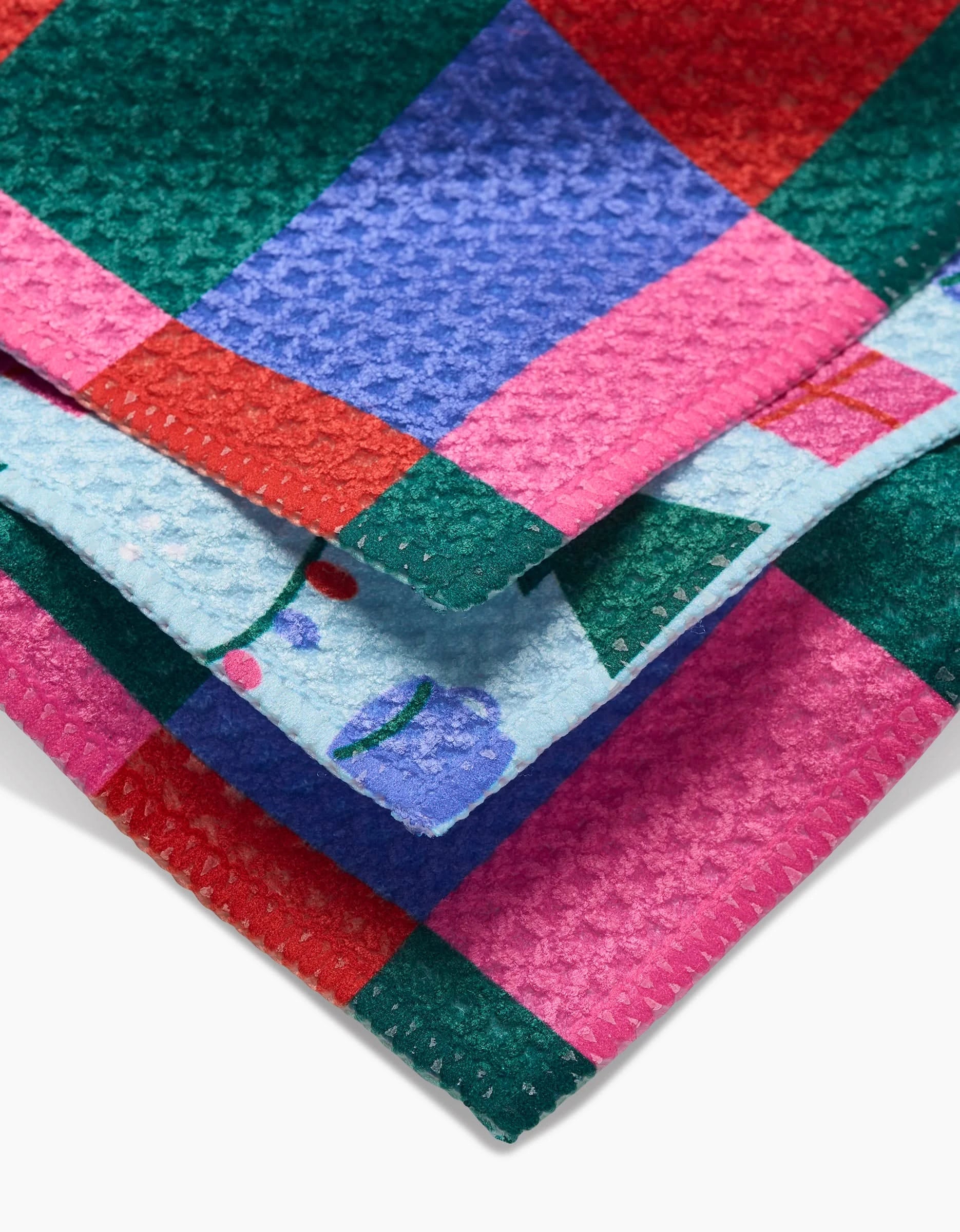 A close-up of Festive Doodles dishcloths by Laura Supnik, featuring colorful checkered patterns in red, green, blue, and pink on recycled material, with cherry and cup designs on a light blue section.