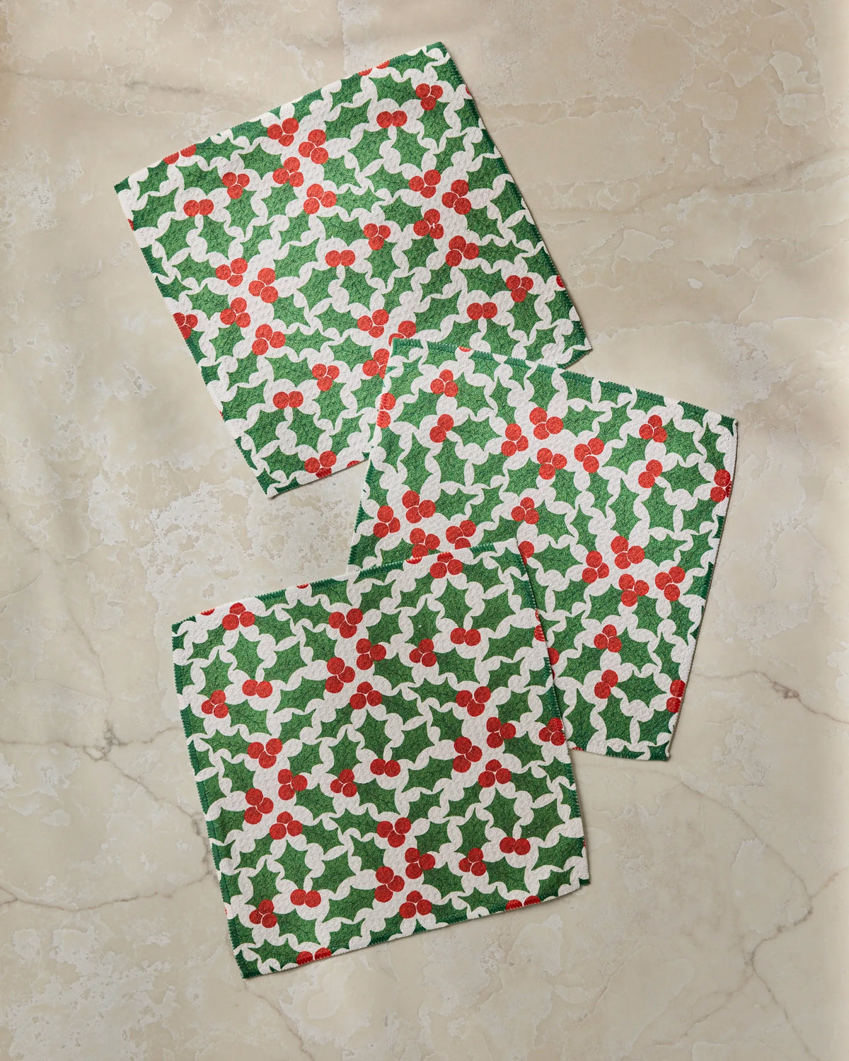 Three Festive Holly square napkins by Geometry, crafted from recycled materials and featuring a green holly and red berry pattern, are arranged on a light beige marble surface.