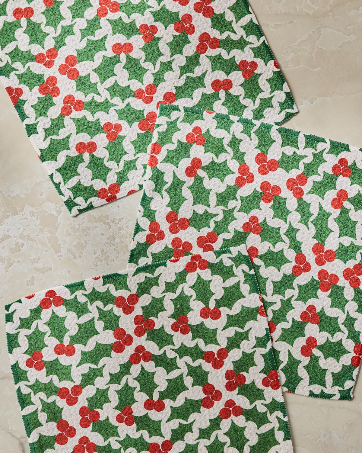 Three square napkins from Geometry’s Festive Holly collection—featuring a lively pattern of green holly leaves and red berries—are arranged on a light, marble-like surface. Thoughtfully crafted from recycled materials, this cheerful and super-absorbent dishcloth set is perfect for everyday use.