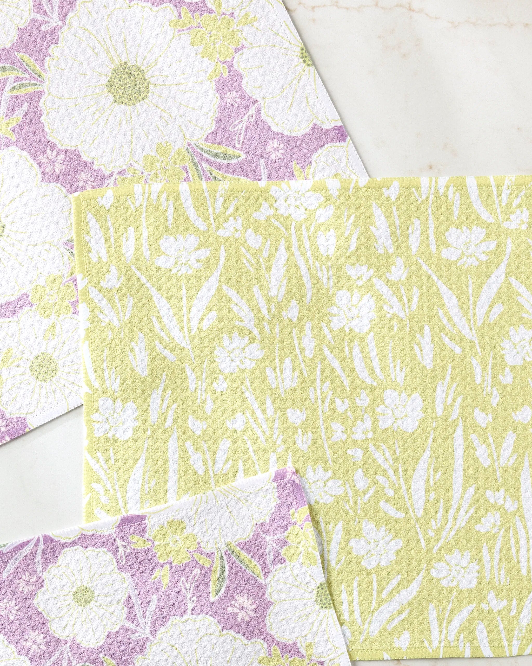 Ashley Celena’s Field of Spring Flowers dishtowels, in yellow and lavender with white floral patterns, are neatly overlapped on a light marble surface.