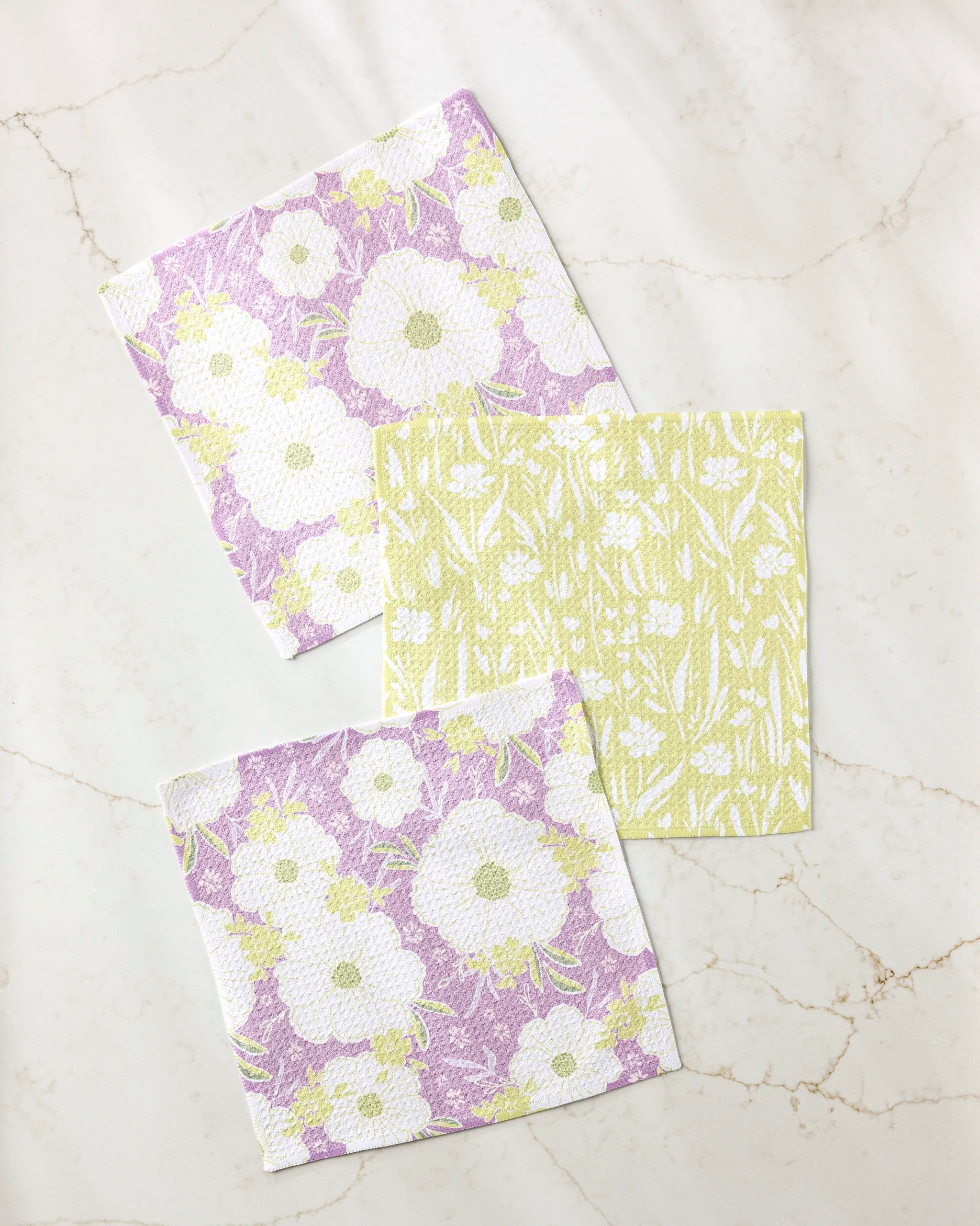 The Ashley Celena "Field of Spring Flowers" dishcloth set includes three square, floral-patterned, super-absorbent dishtowels—two in purple, white, and yellow, and one in yellow and white—displayed on a light marble surface.