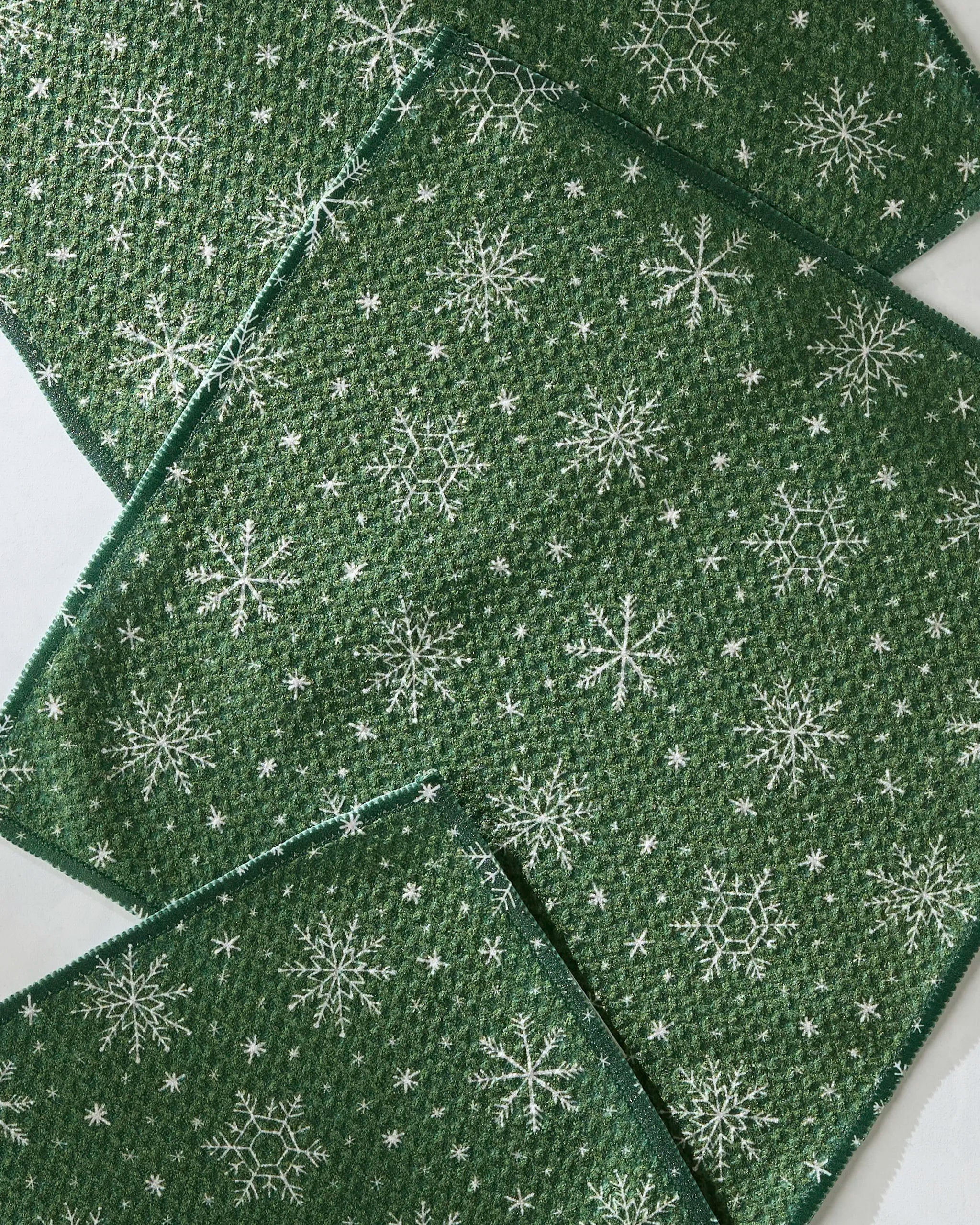 Four green Firlight napkins by Geometry, featuring a textured weave and different white snowflake patterns, overlap on a light surface. Crafted from recycled materials.