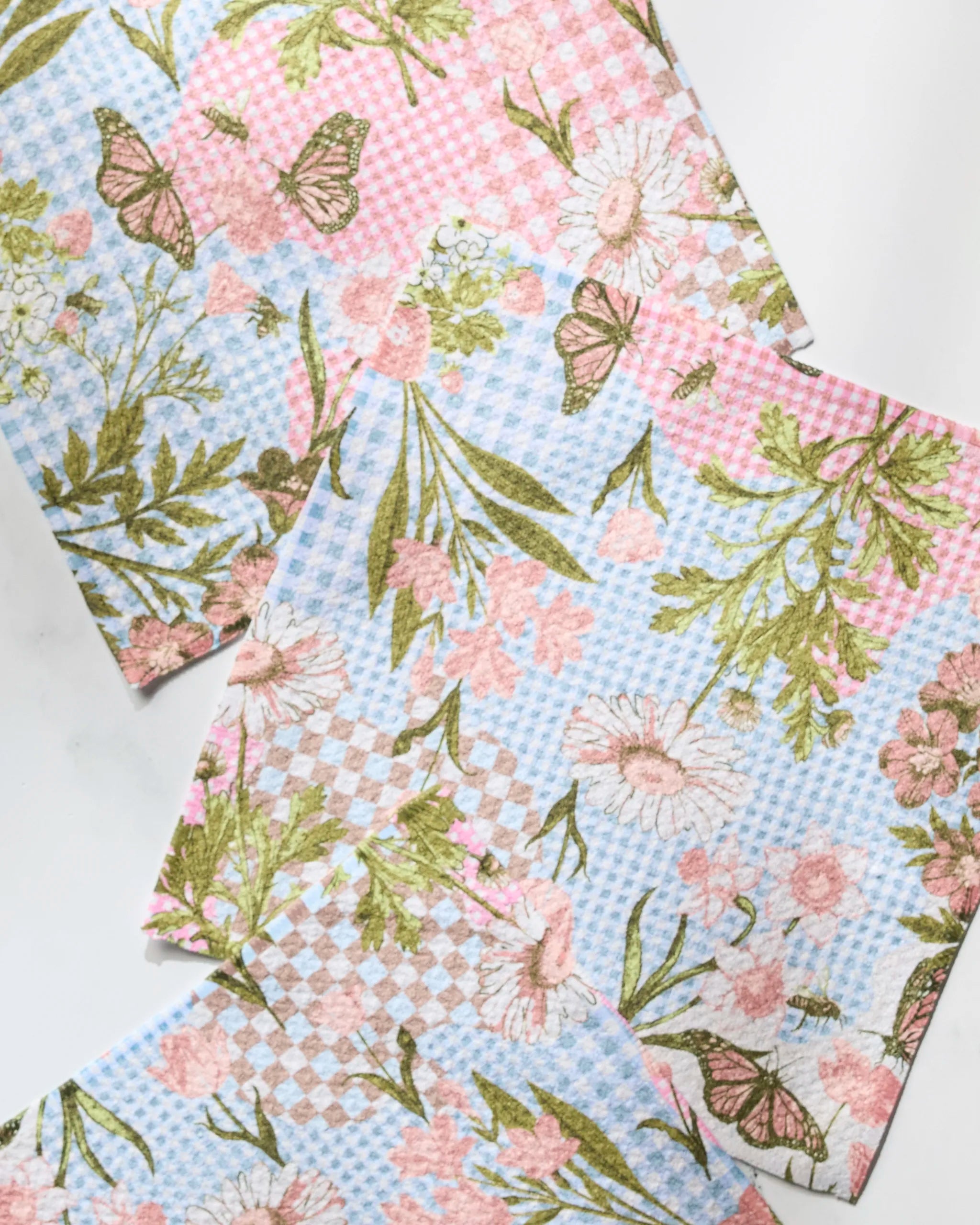 Three "Floral Collage" paper napkins by Geometry, featuring pink and blue checkered backgrounds with floral patterns, butterflies, and green leaves, are arranged on a white surface—perfect to pair with recycled kitchen towels.