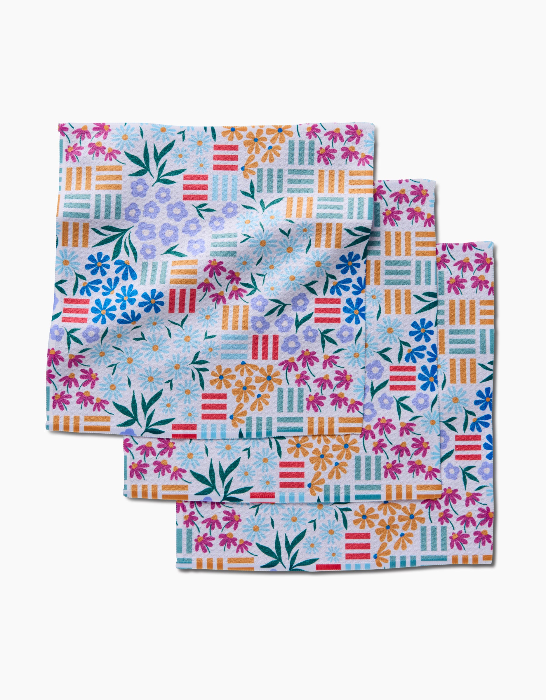 Three Floral Garden napkins by Charlie Rowan Designs—made from recycled material—feature orange, pink, blue, and purple flowers, green leaves, and a geometric grid on white, arranged in an overlapping stack.