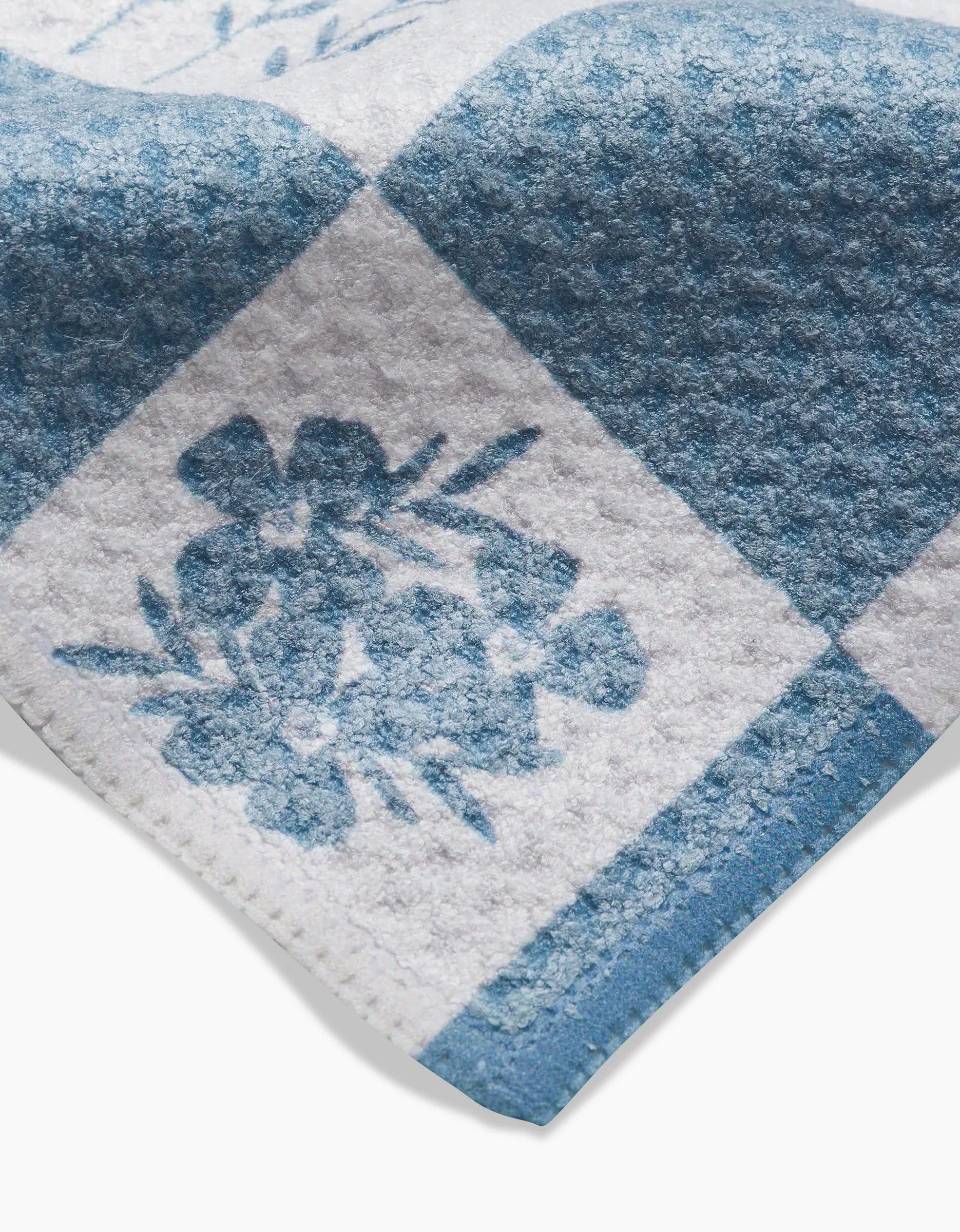 A close-up of the Floral Patchwork towel by Charlie Rowan Designs shows its blue and white checks with a floral square. Made from soft, textured Geoweave fabric.