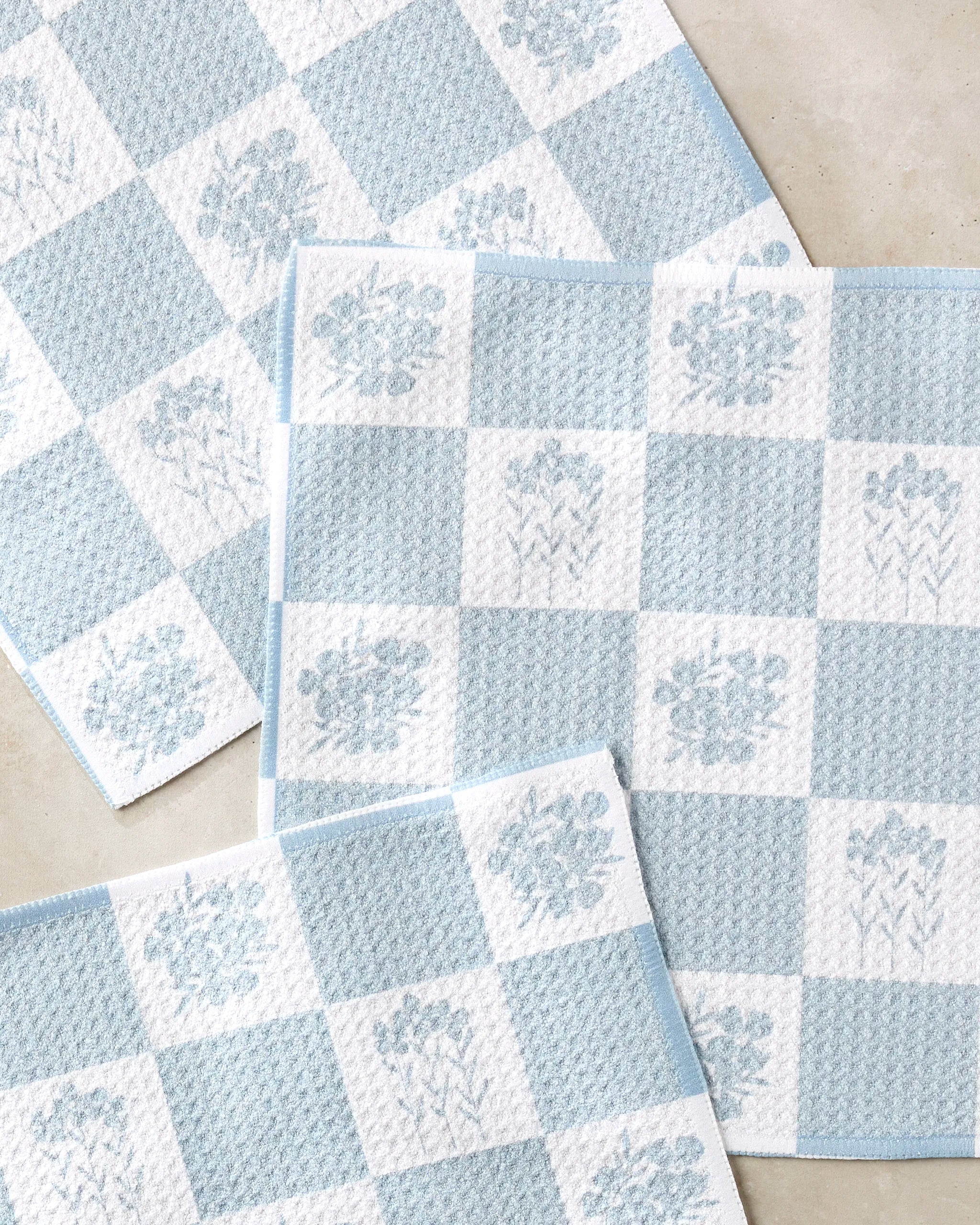 Four Charlie Rowan Designs "Floral Patchwork" dish towels, featuring blue and white checks with subtle florals and made from recycled materials, are slightly overlapped on a light surface.
