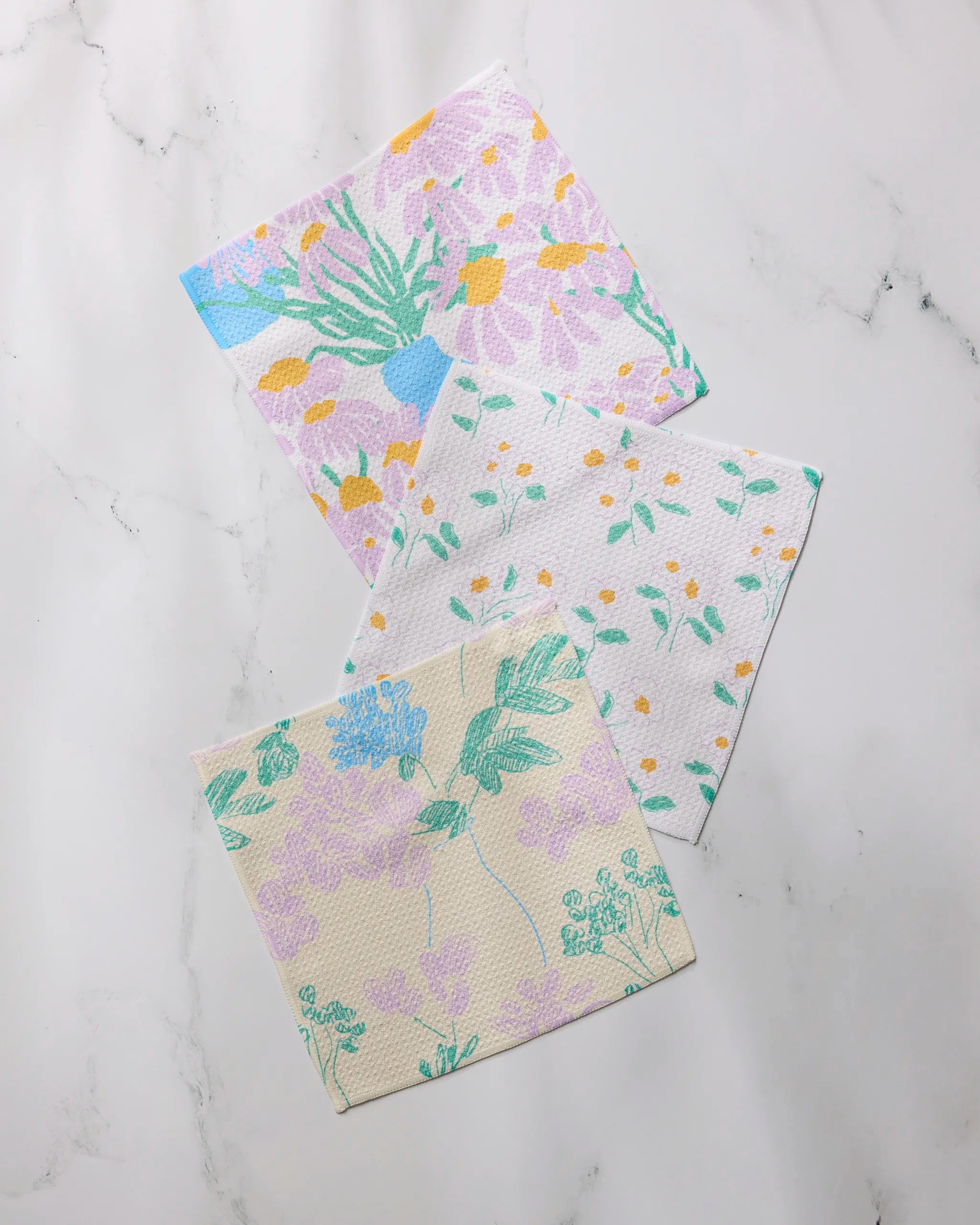 Geometry’s Florals for Mom dishcloth set includes three square cloths featuring unique pastel botanical designs in green, blue, purple, and yellow, all crafted from recycled materials.