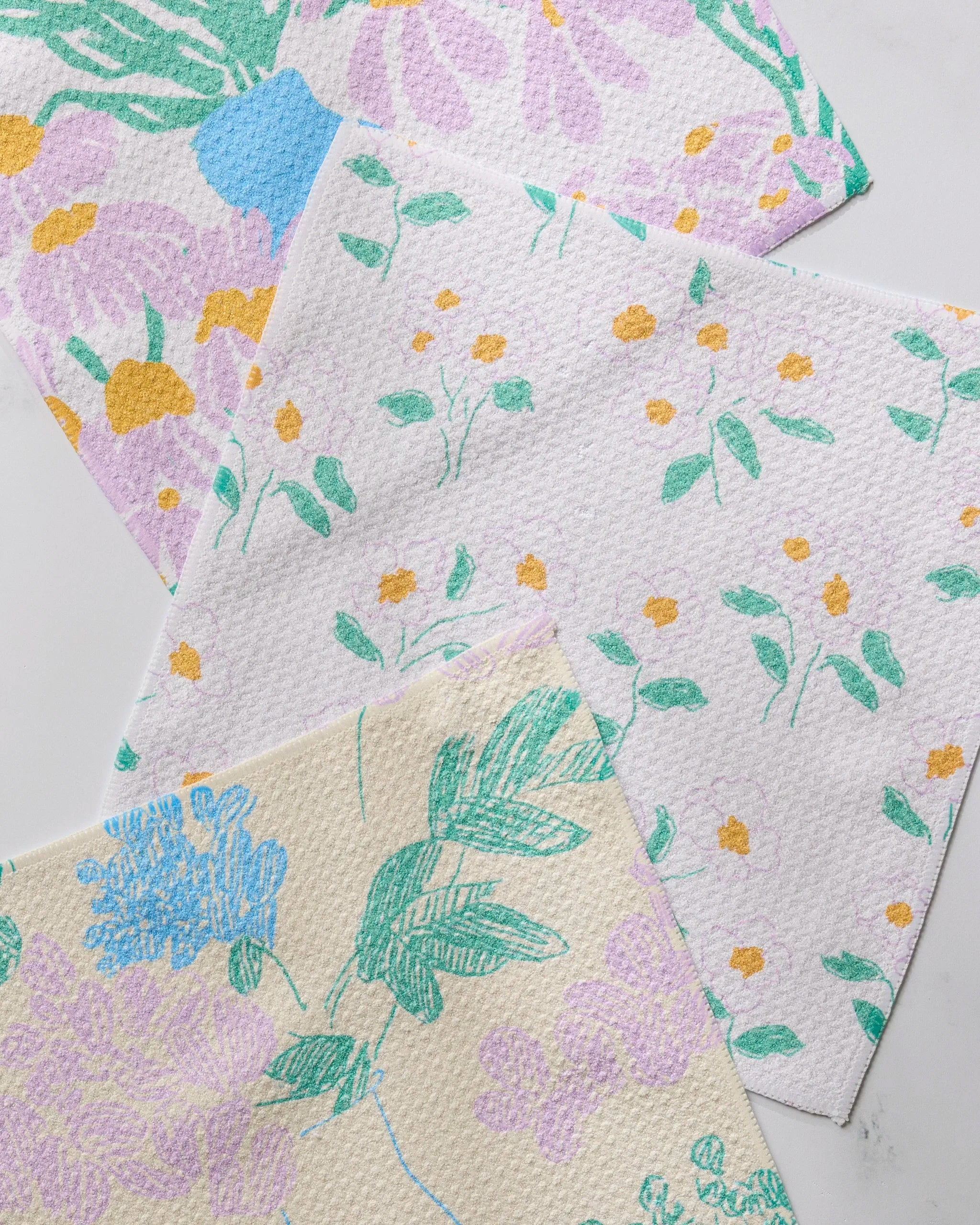 Three "Florals for Mom" cloth napkins by Geometry, each with a unique pastel floral pattern and green leaves, are displayed on a white surface. This eco-friendly set is made from recycled materials.