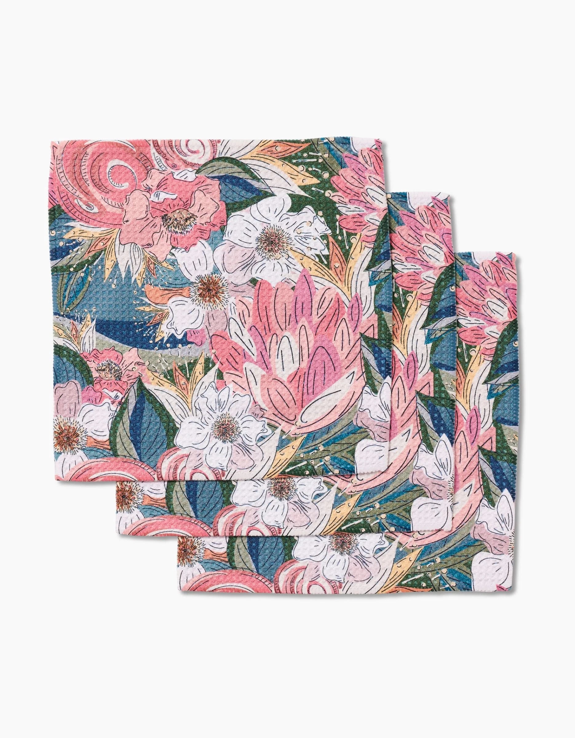 Three folded Flower Explosion napkins by Second Nature Studio, featuring a vibrant pink, white, and orange floral pattern with green leaves on blue Geoweave fabric.