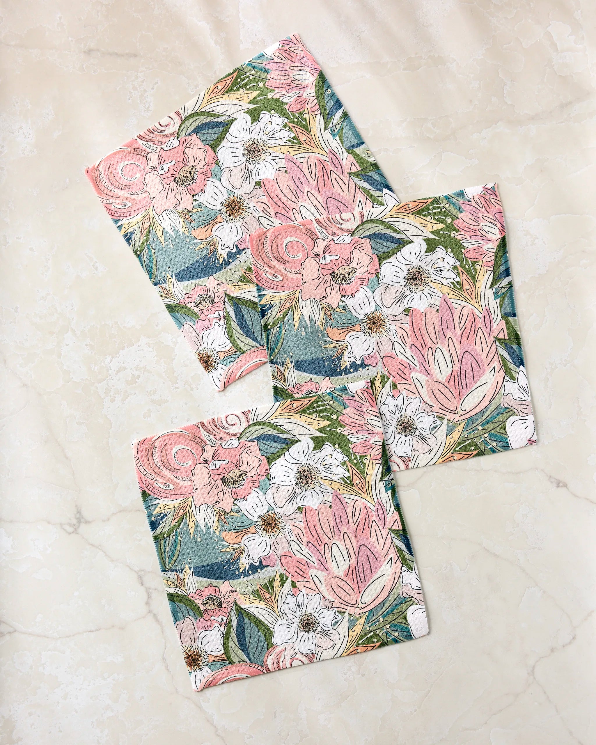 Three Flower Explosion reusable fabric wraps by Second Nature Studio, made from recycled materials, feature a pink, white, and green floral pattern and are arranged overlapping on a light marble surface.