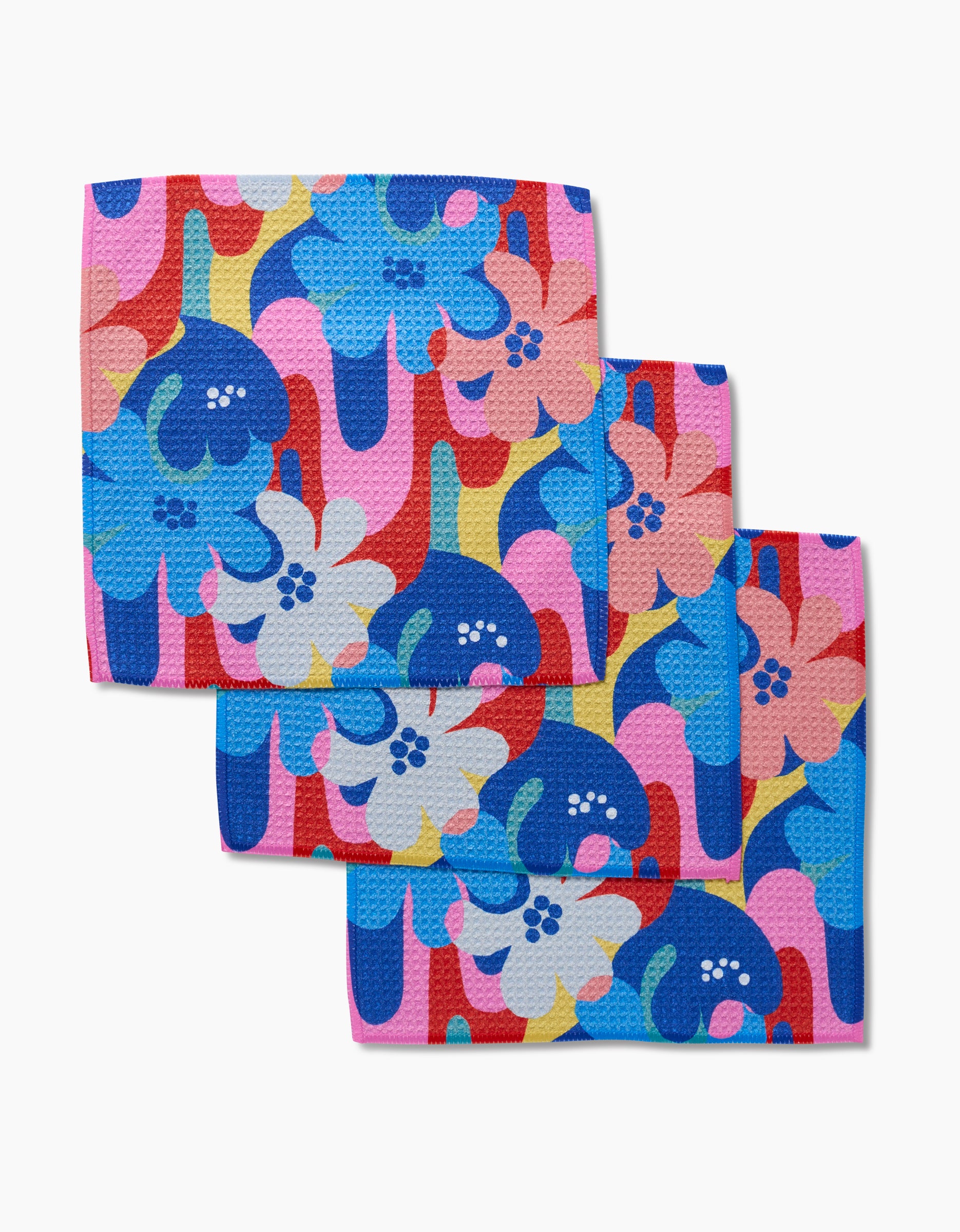 Three Flower Powers dishcloths by Masha Volnova, featuring a vibrant floral pattern, are stacked on a white background. Each is crafted from Geoweave fabric in blue, pink, red, white, and yellow.