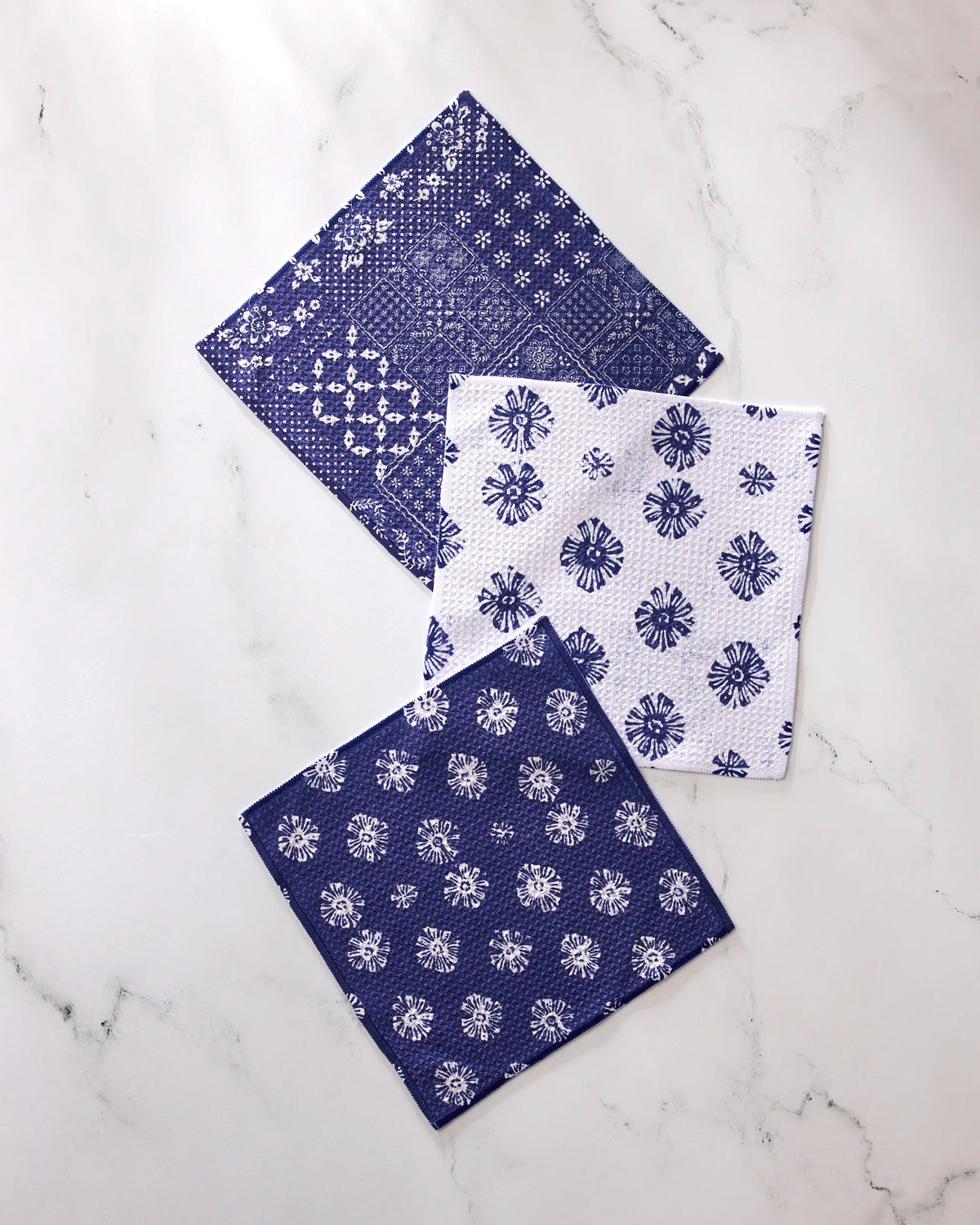 Three Flowing Ocean napkins by Geometry are displayed on white marble: two in navy with white patterns, and one in white with a blue floral design.