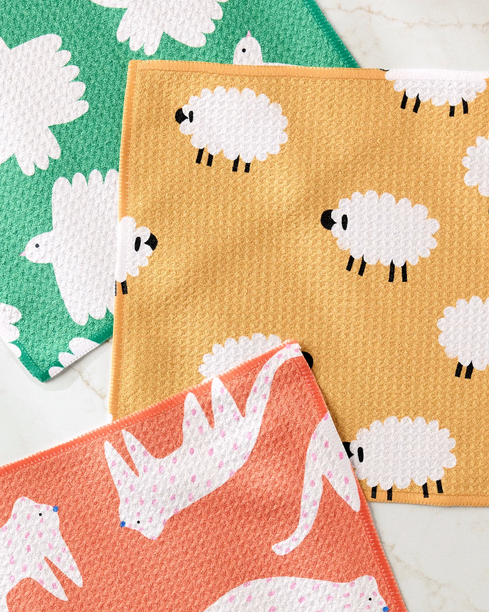 The Fluff and Feathers dishcloth set by Jennifer Bouron includes three recycled, super-absorbent towels—green and yellow with white sheep, and coral with playful cat-like animals accented by blue or pink dots.