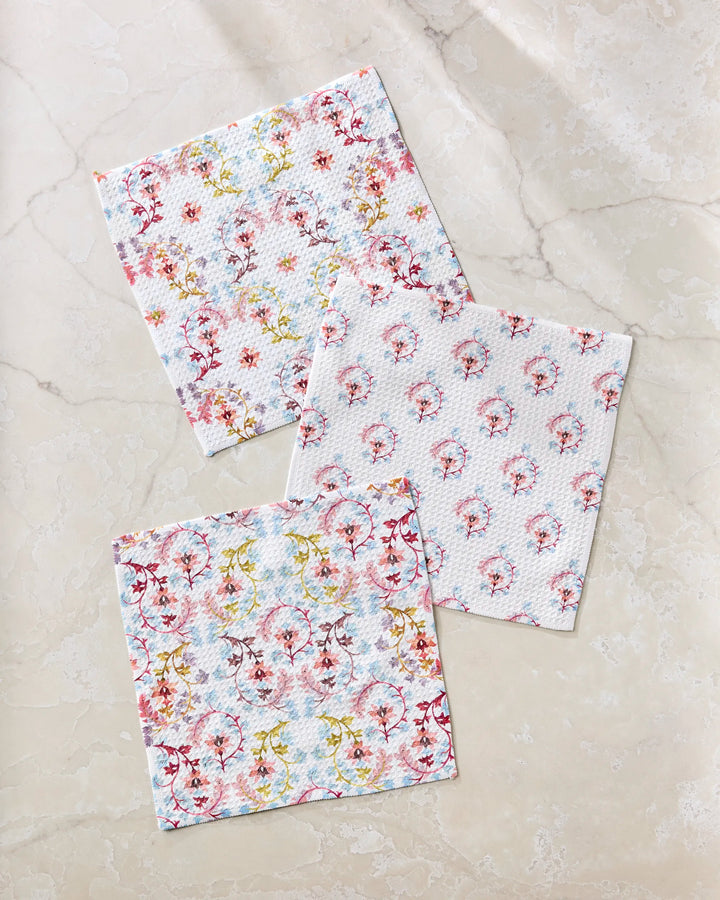 The Folk Medallion dishcloth set by Maayan Alper-Swan includes three square cloths with pink, red, and yellow pastel floral patterns, displayed on a beige marble surface. Made from recycled materials for eco-friendly style.