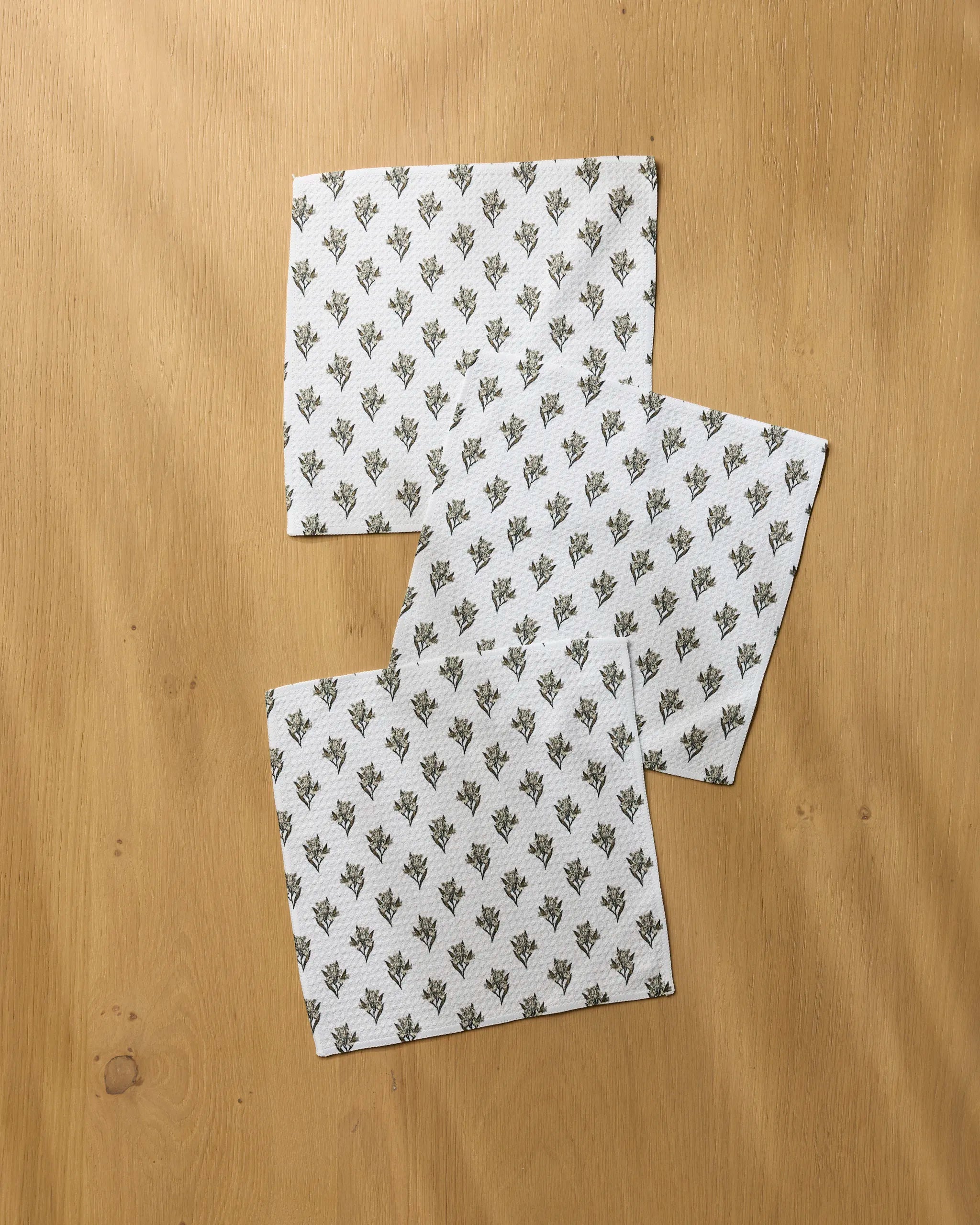 Three Foraged napkins by Geometry, featuring a subtle black floral pattern, are arranged diagonally and slightly overlapping on a light wooden surface, creating an elegant display reminiscent of recycled kitchen towels.