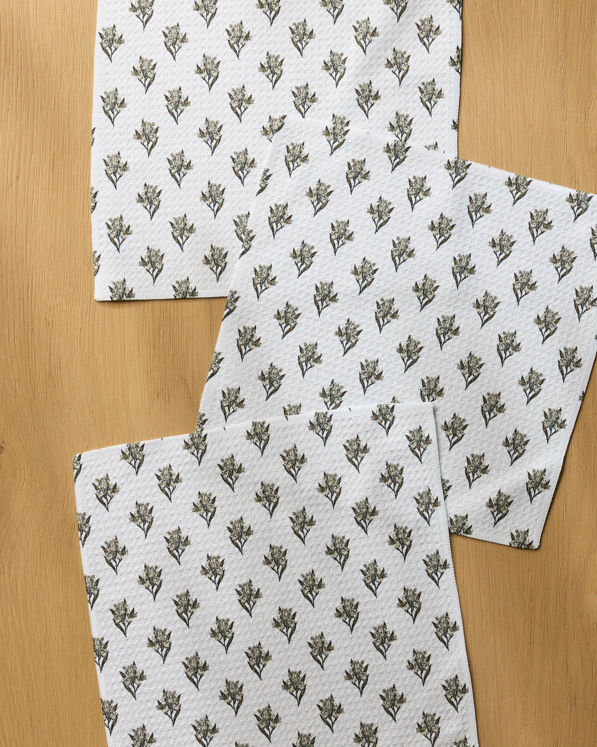 Three "Foraged" kitchen towels by Geometry, featuring a small repeating floral pattern on white recycled fabric, are laid out slightly overlapping on a light wooden surface.