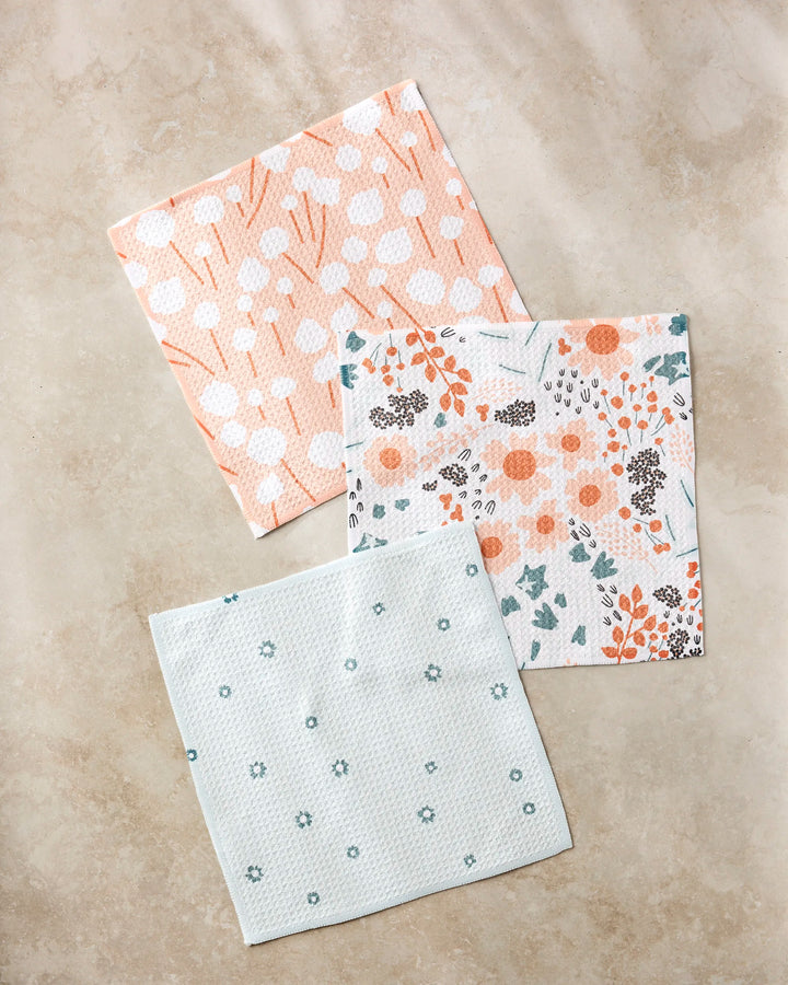 The Forest Florals set by Coit Creative features three reusable cloths—orange dots and lines, orange-blue florals, and blue flowers—arranged on a beige surface for a charming, eco-friendly dishcloth set.