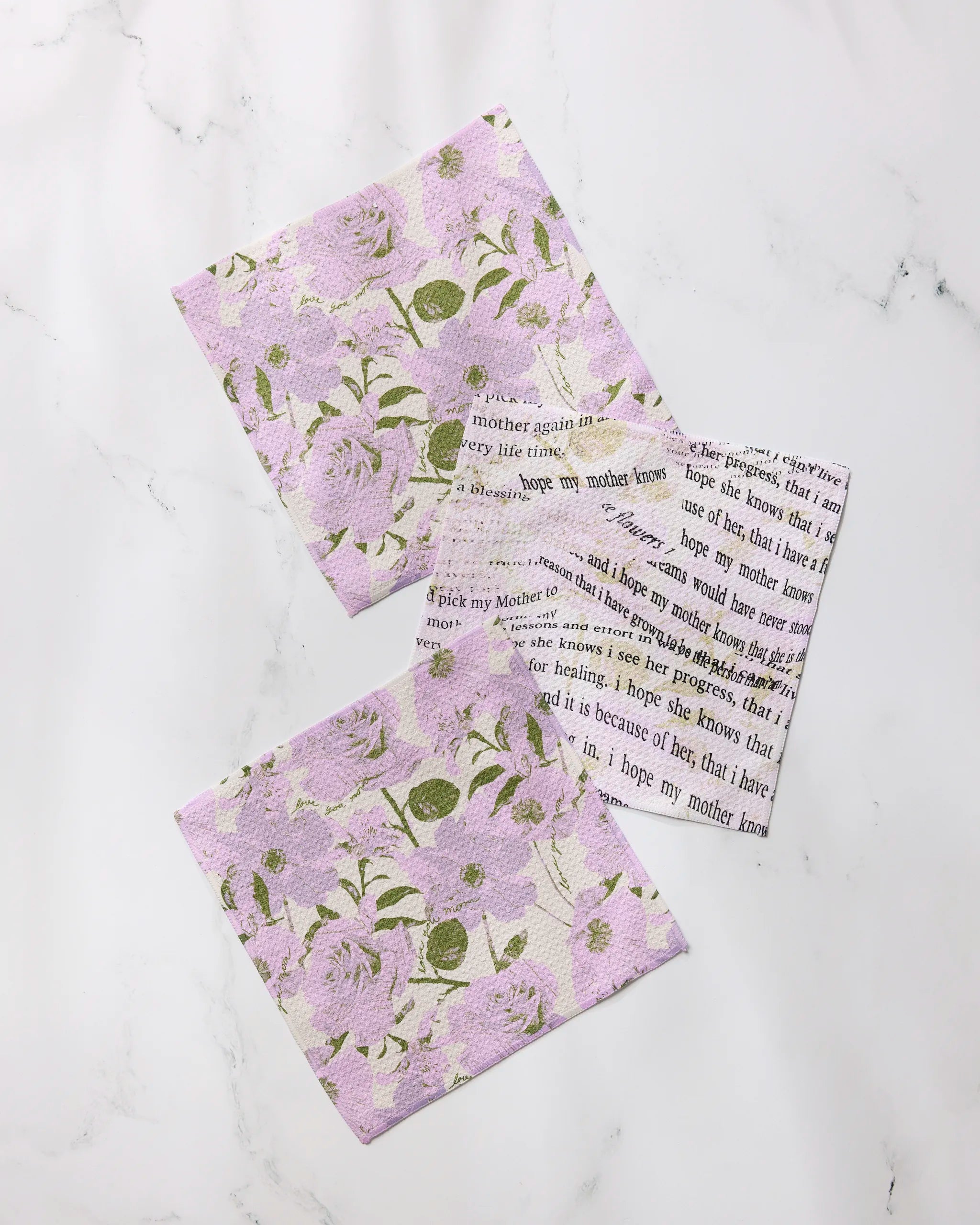 Three fabric squares from Geometry’s Forever Mom dishcloth set are displayed on white marble; two show a purple floral pattern with green leaves, and one features black text on a white background.