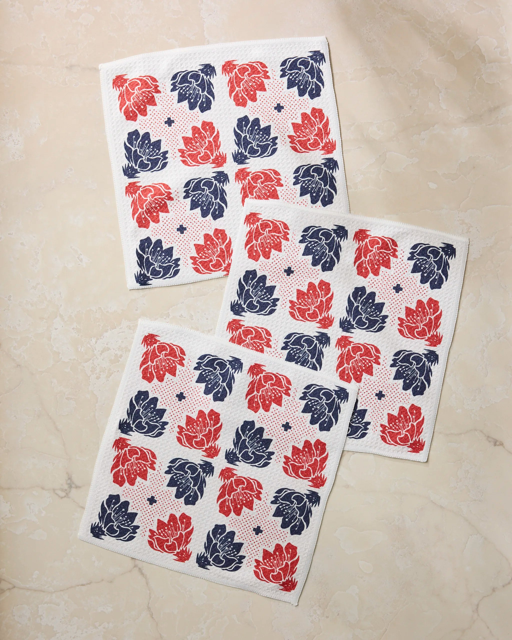 Three Founding Floral square napkins by Geometry, made from recycled material, display a red and navy blue floral pattern on a light beige marble surface.
