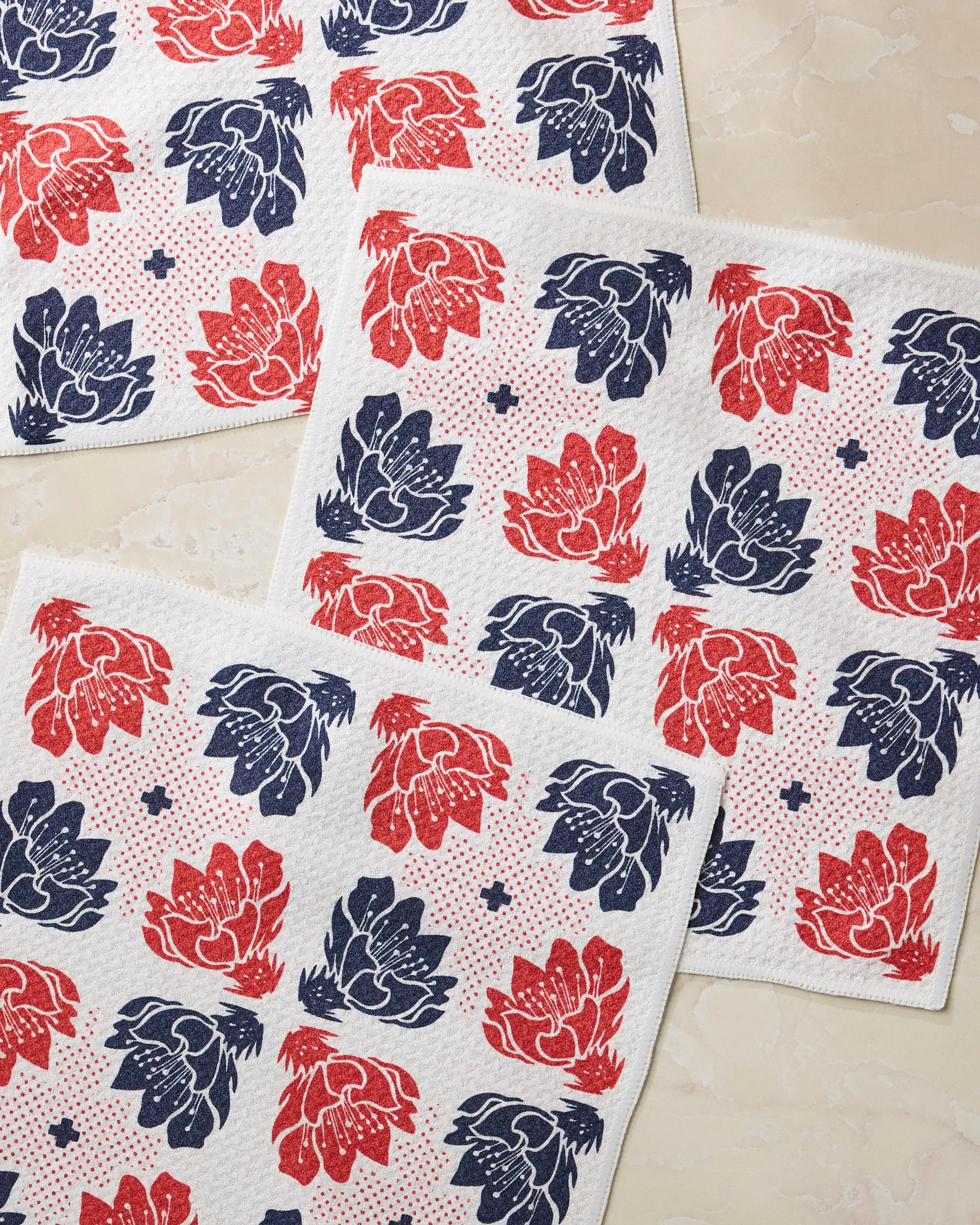 Geometry’s Founding Floral set features three textured napkins with red and blue floral patterns and small blue plus signs, ideal as absorbent kitchen towels or reusable dishcloths, arranged on a light marbled surface.