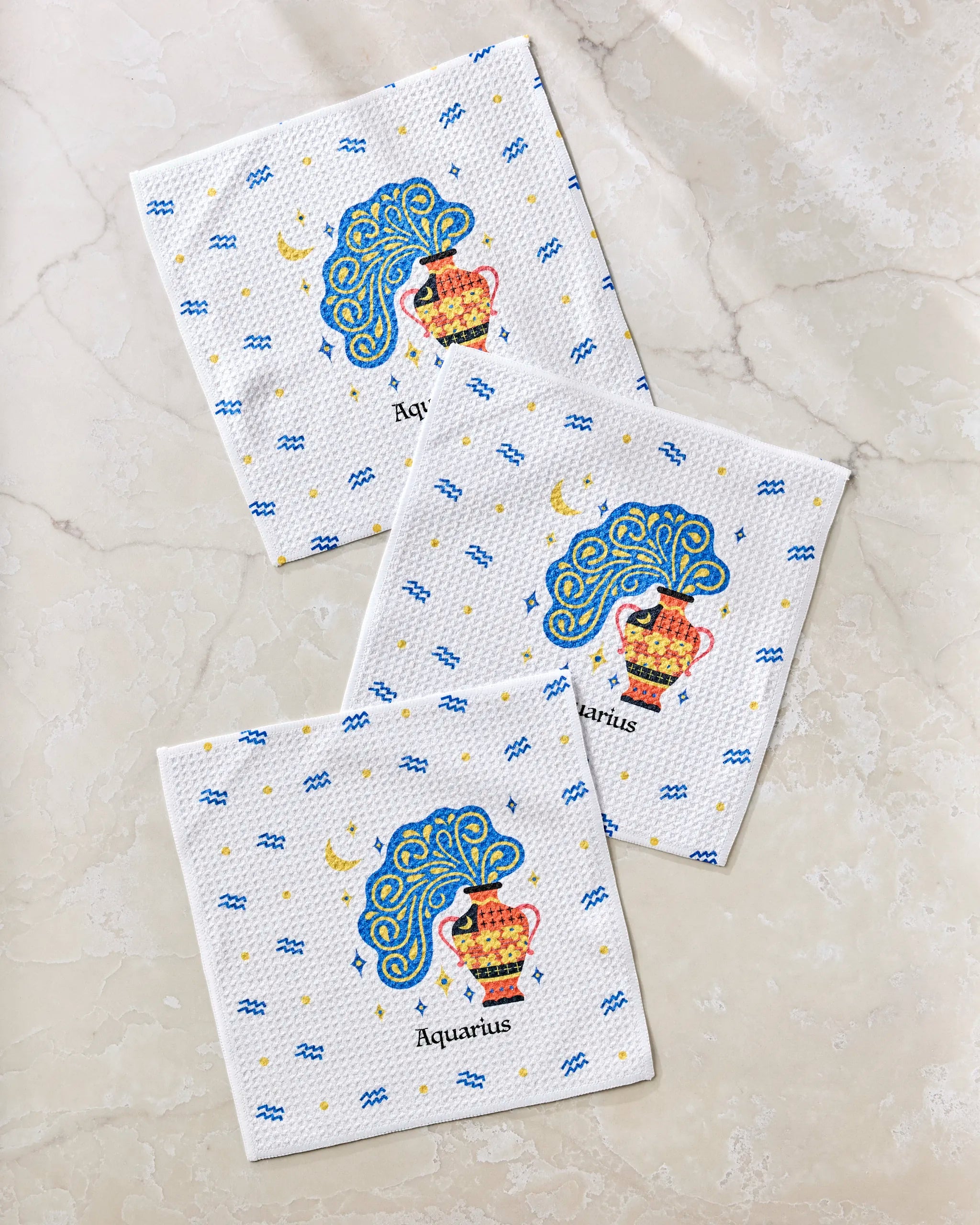 Three "Free Aquarius" paper napkins by Heather Zhou, featuring a colorful Aquarius zodiac design with a water jug and blue waves, made from recycled materials and arranged on a light marble surface.