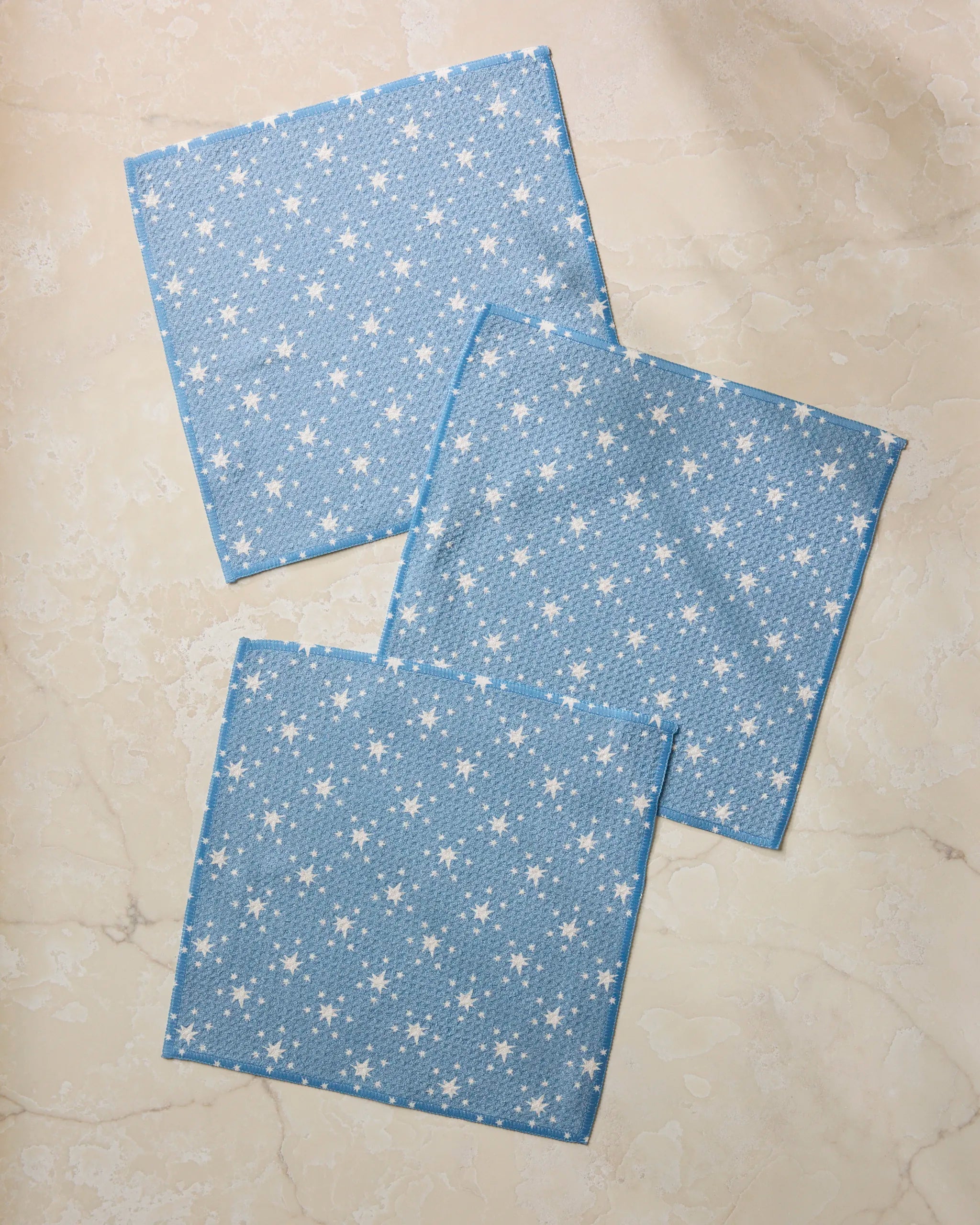 Three Geometry Freedom Blue napkins, made from recycled materials and featuring a small white star pattern, are laid out overlapping on a light marble surface.