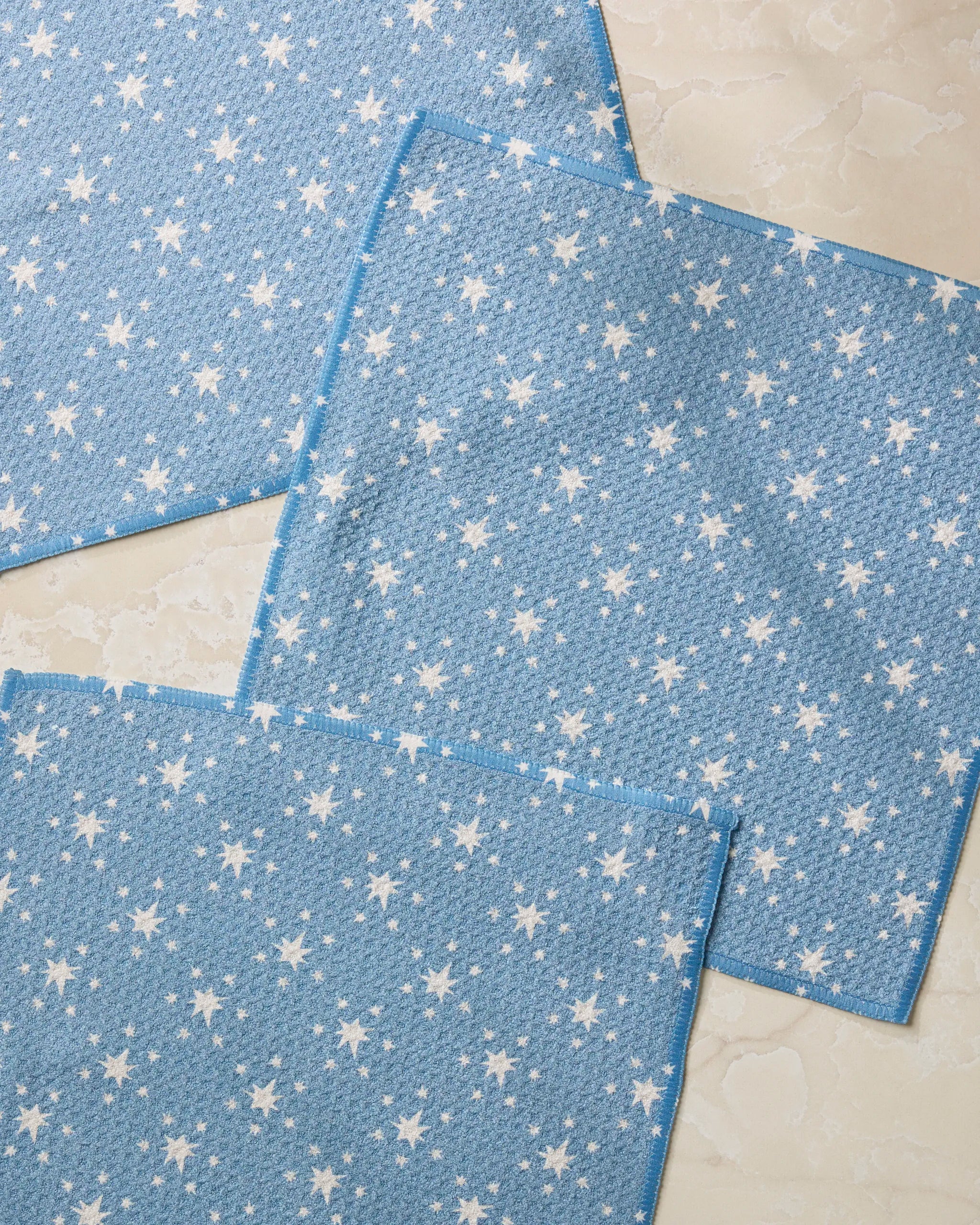 Three Freedom Blue fabric squares by Geometry, featuring a white star pattern and crafted from recycled materials, are arranged overlapping each other on a light marbled surface.