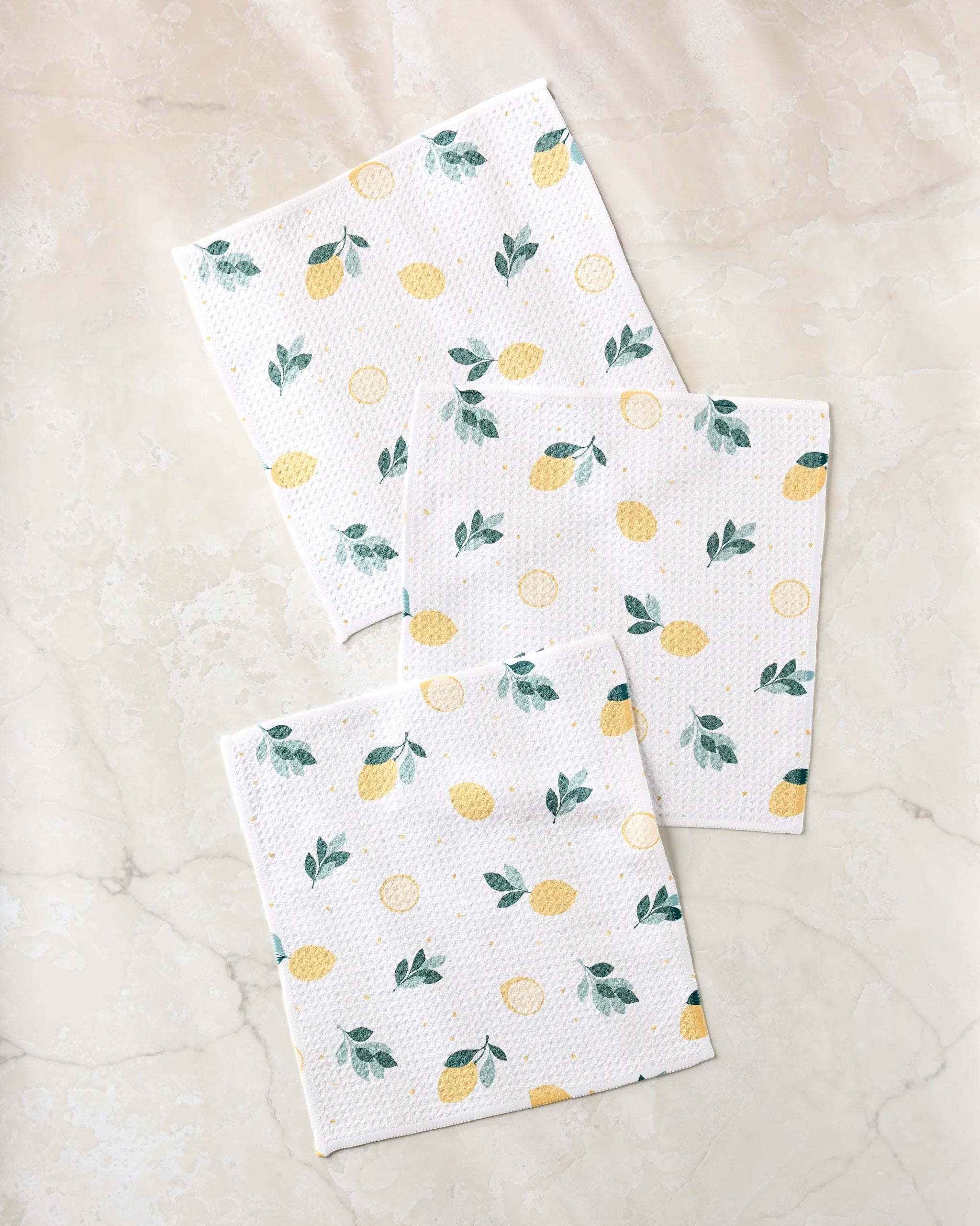 The Sara Mari "Freshly Picked" dishcloth set features three white cloths with yellow lemons and green leaves, laid on marble. Crafted from recycled materials, they're a stylish and practical eco-friendly cleaning choice.