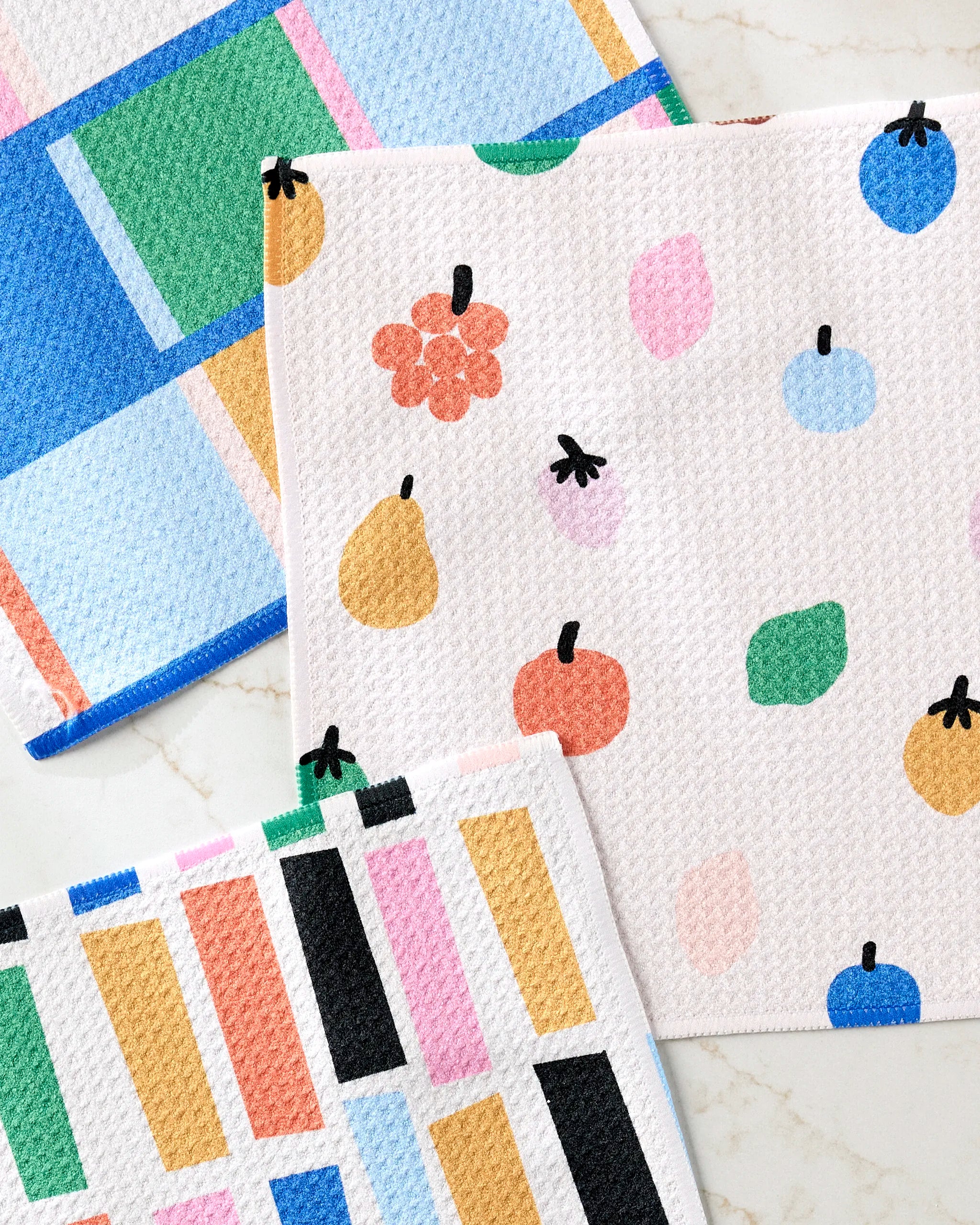 Three Fruit Basket dishcloths by Jennifer Bouron, made from recycled materials, are displayed on light marble. Each features a playful design: geometric patterns, abstract dots, and fruit illustrations in pastel and bold colors.