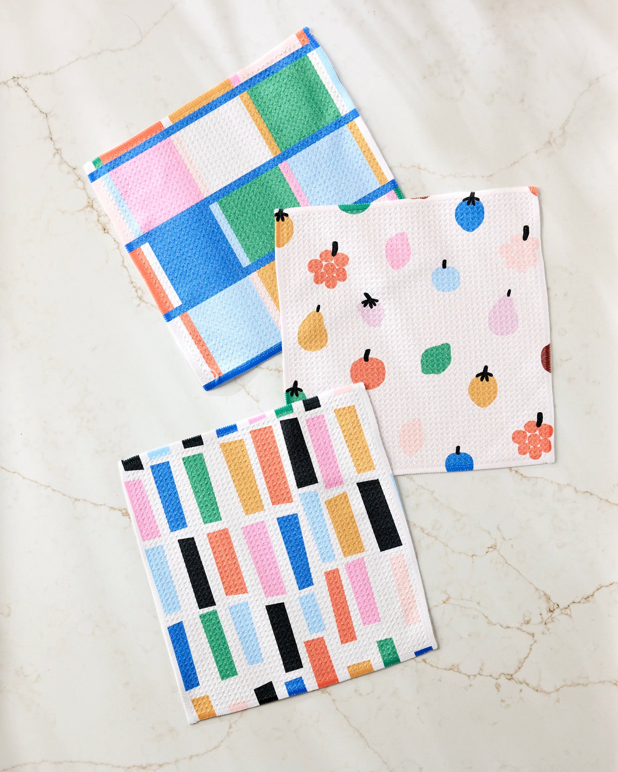 Three colorful Fruit Basket dishcloths by Jennifer Bouron rest on marble: one sports blue, green, pink, and beige checks; another has a fruit print; the third features multicolored vertical rectangles.