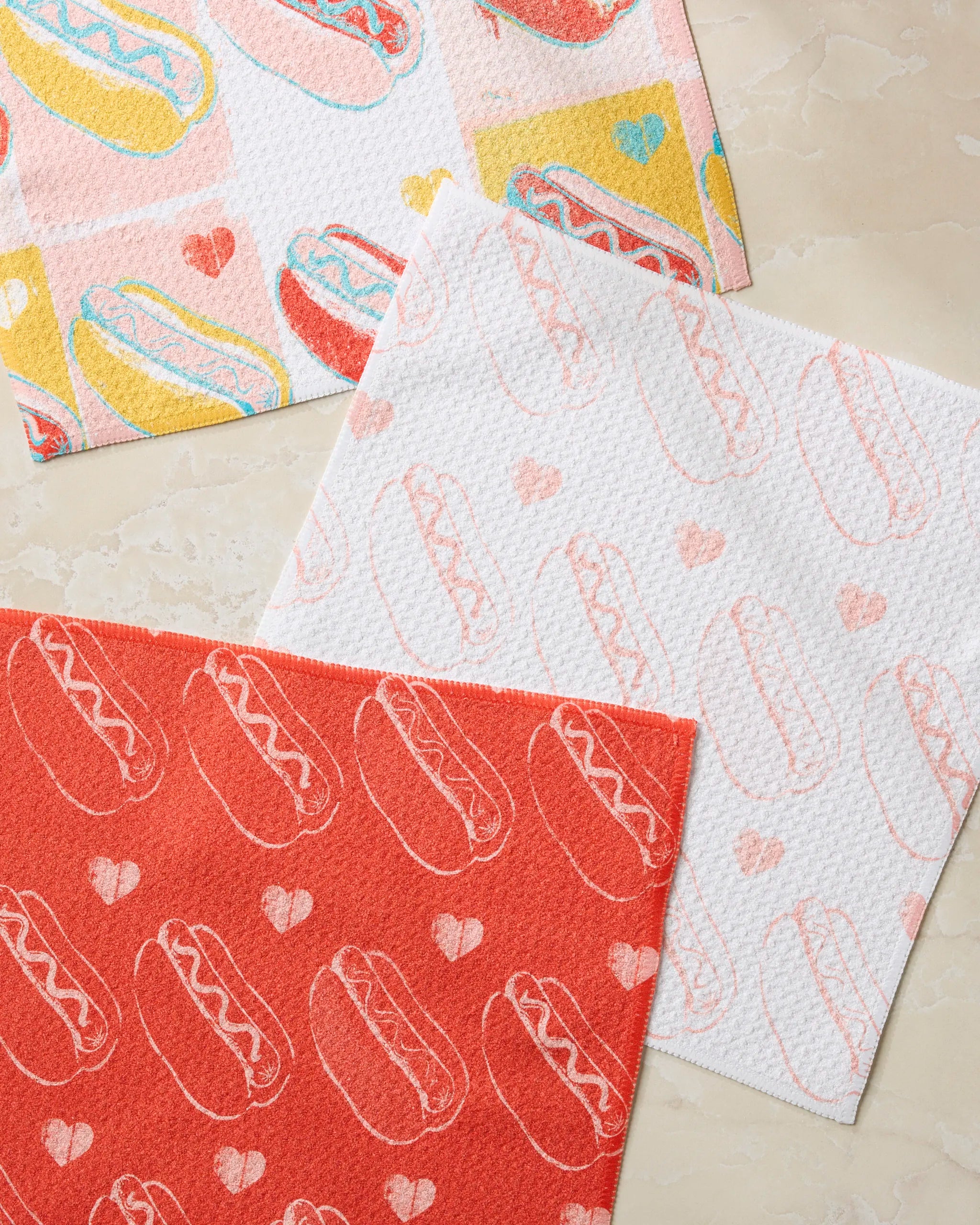 Three absorbent Geometry Fun Franks dishcloths with hot dog and heart designs, in red, white, and multicolor, are displayed on a light marble surface—perfect for adding a playful touch to your kitchen.