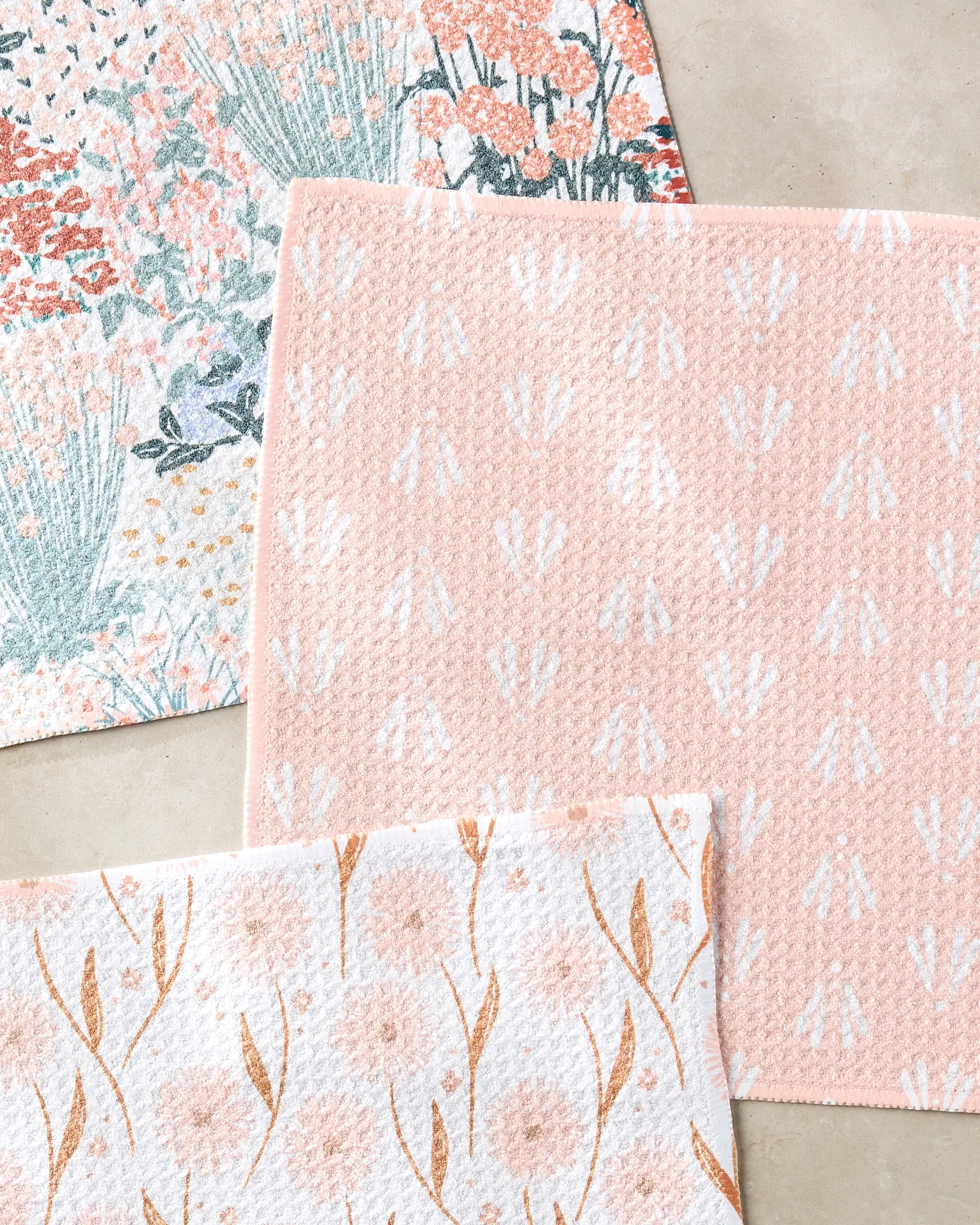 Three pastel-hued Garden Bloom bath mats by Hufton Studio, crafted from recycled materials and featuring pink, white, and green floral and botanical motifs, are arranged on a light beige floor.