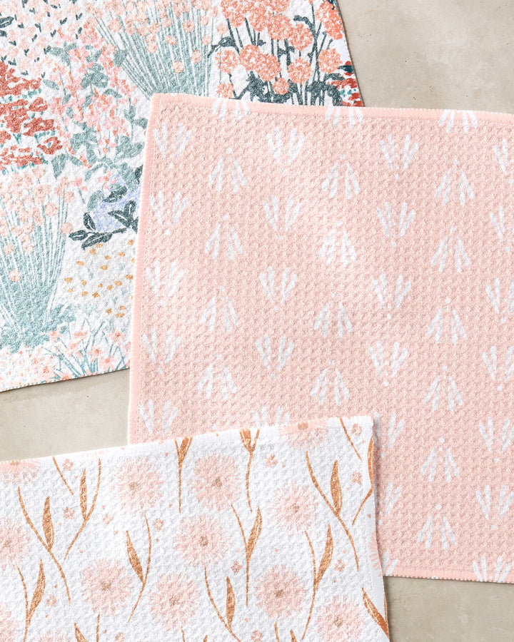 Three pastel-hued Garden Bloom bath mats by Hufton Studio, crafted from recycled materials and featuring pink, white, and green floral and botanical motifs, are arranged on a light beige floor.