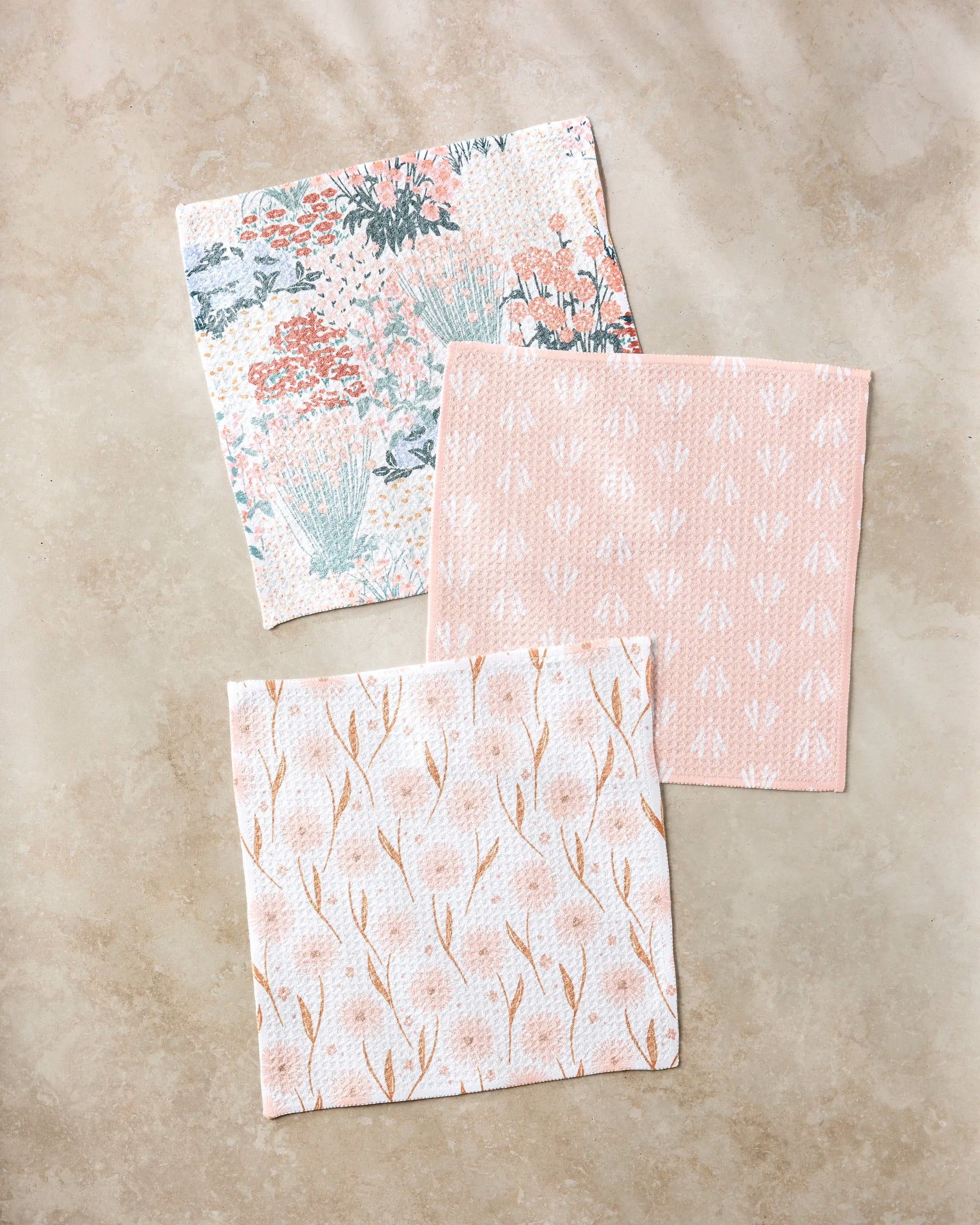 Three square cloths from Hufton Studio’s Garden Bloom set—super-absorbent and decorated with pastel floral, arrow, and abstract branch designs—are displayed on a light beige marble surface.