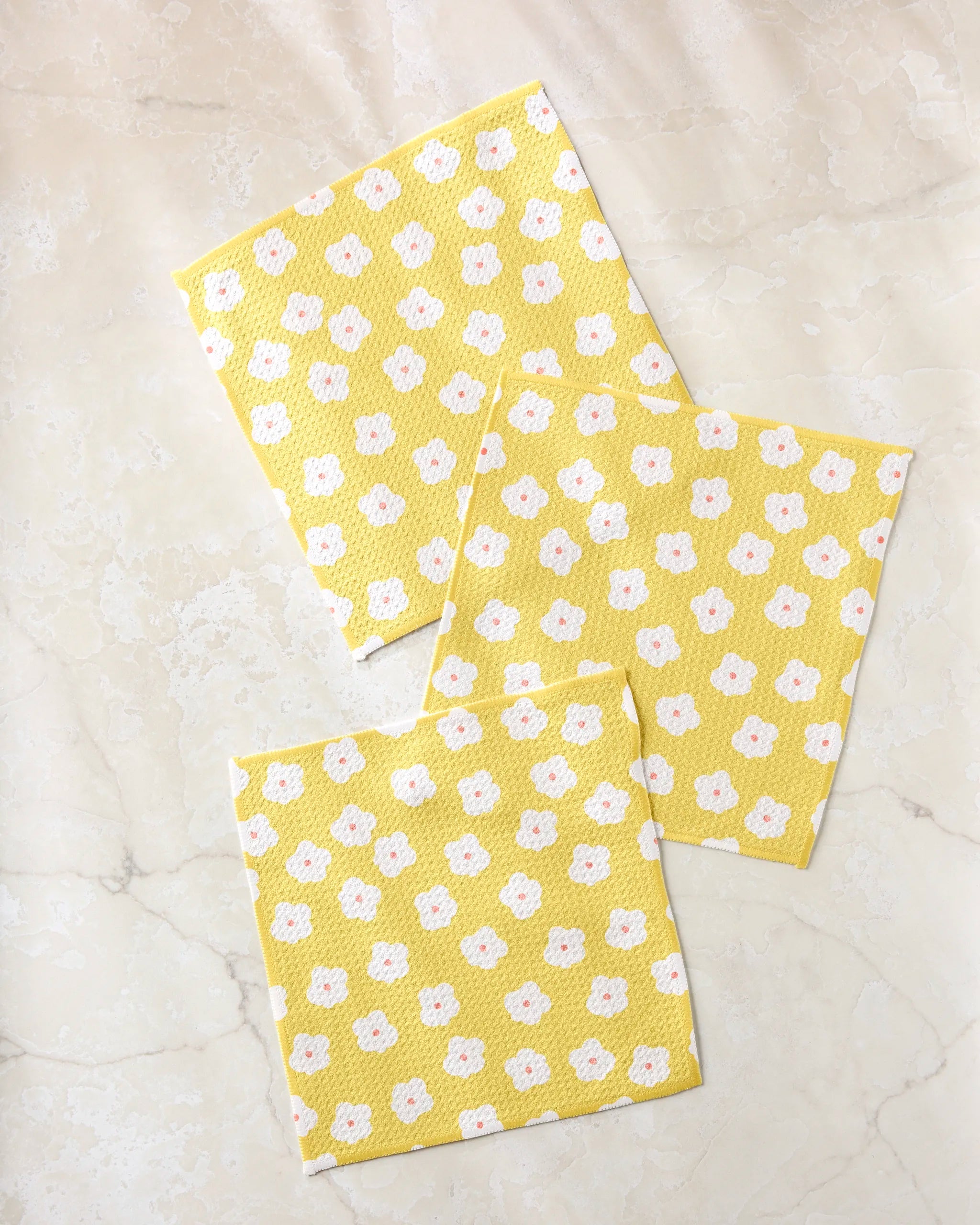 Three Garden Crush cloths by Sara Mari, featuring a white and orange floral pattern on yellow and made from recycled materials, are displayed on a light beige marbled surface.