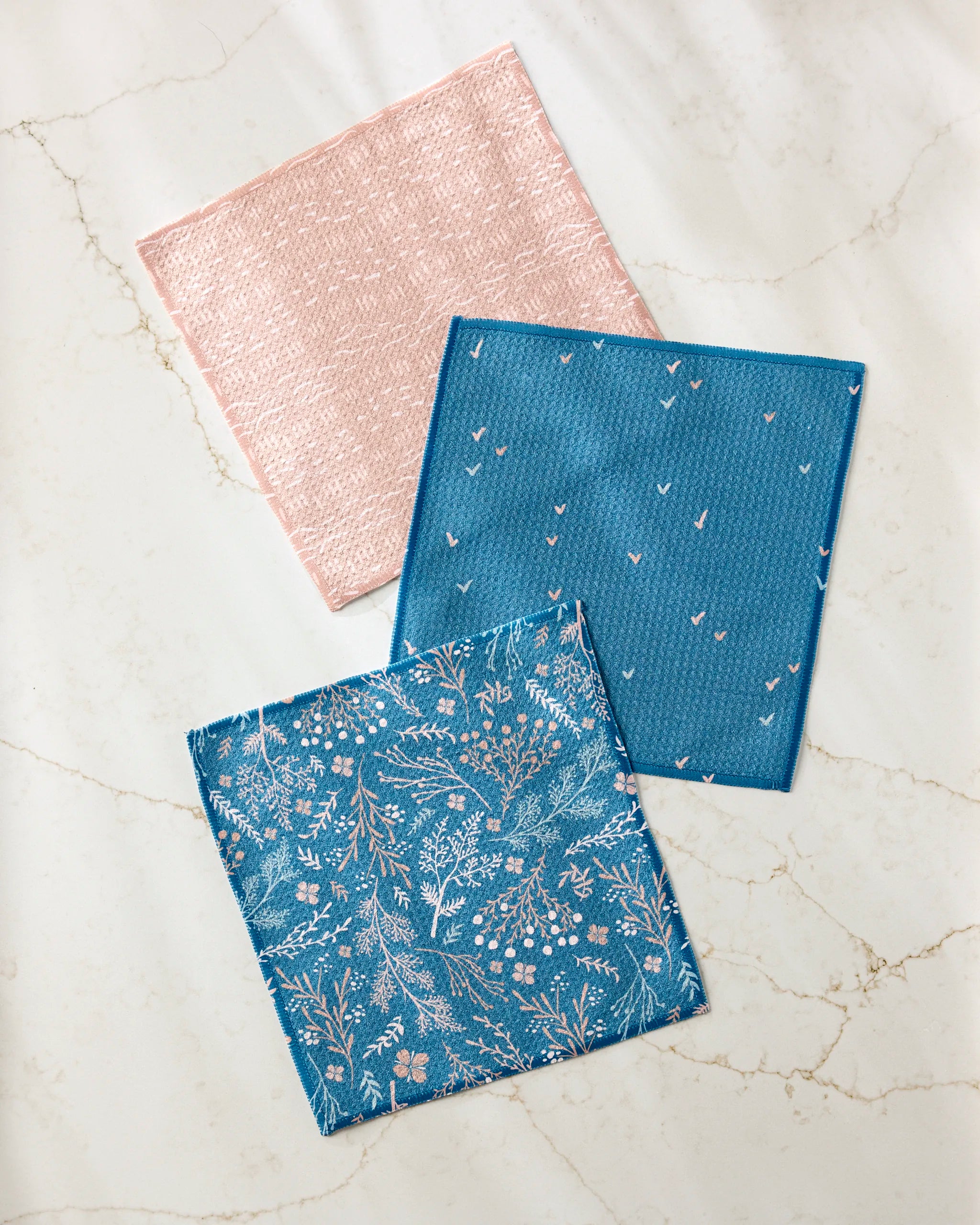 Three "Garden Seeds" cloth napkins by Danelys Sidron, made from recycled materials, are displayed on a light marble surface: one pink, one blue with white arrows, and one blue with a white floral design.