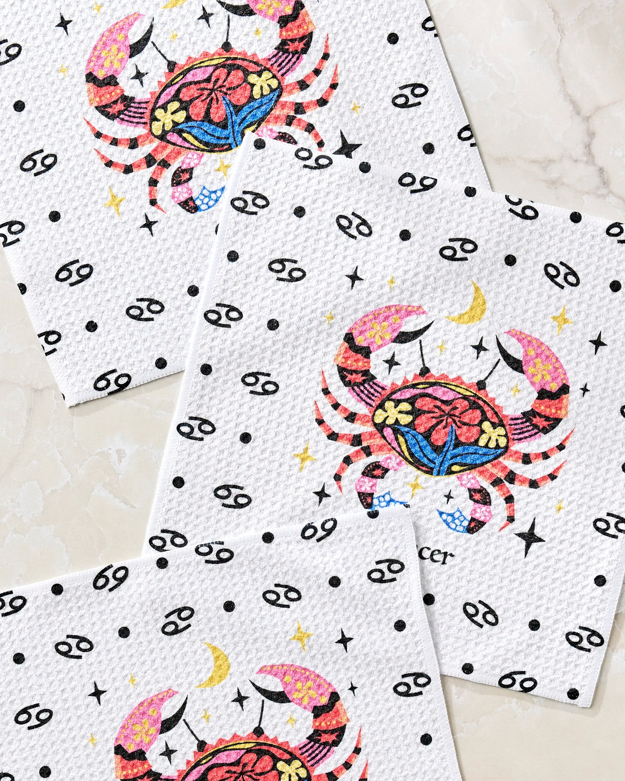 Three white "Gentle Cancer" napkins by Heather Zhou, made from recycled materials, feature a colorful crab, crescent moons, stars, and the Cancer zodiac symbol in black on a light marble surface.