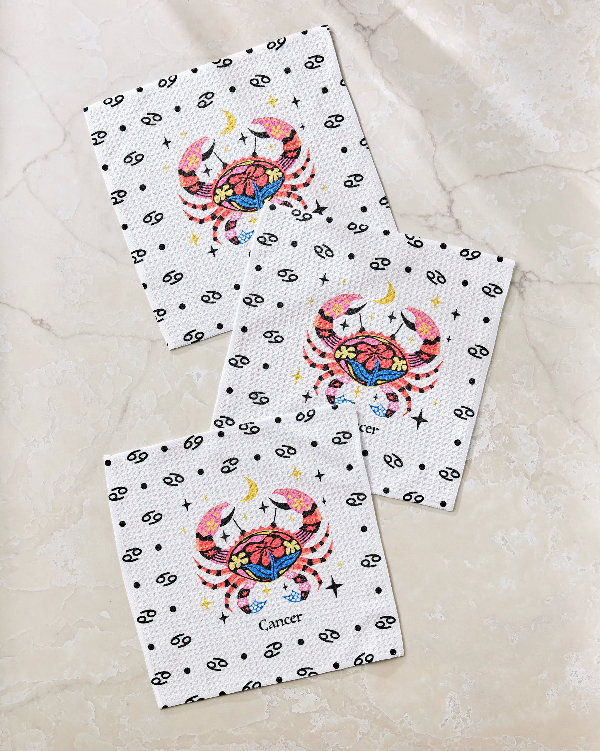 The Gentle Cancer dishcloth set by Heather Zhou includes three white cloths with colorful crab and Cancer zodiac designs, crafted from recycled materials. One features the word "Cancer," all arranged on a light marble surface.