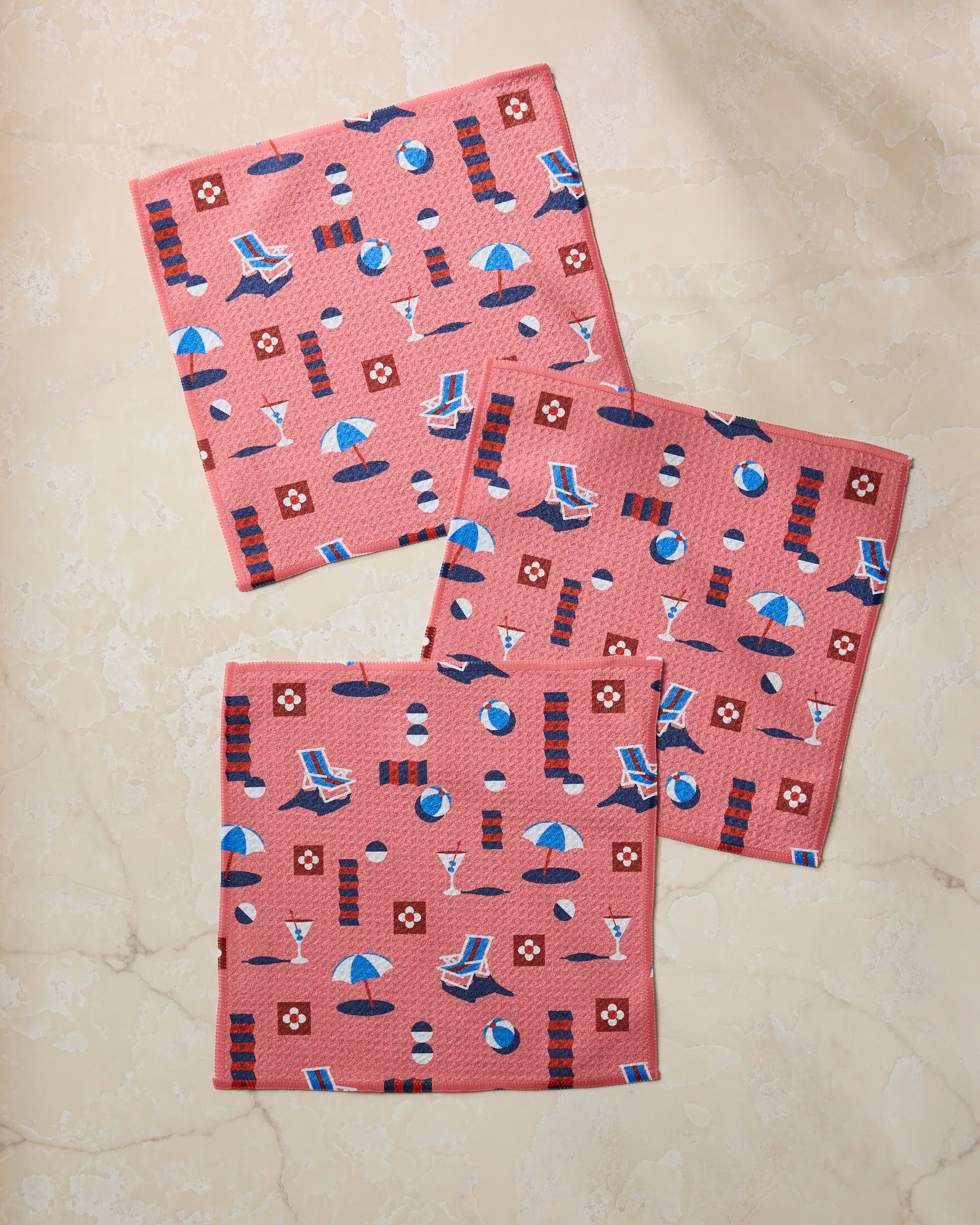 Three square pink Geometry "Geo Beach Day" towels made from recycled materials feature a beach-themed pattern with umbrellas, lounge chairs, beach balls, drinks, and sunscreen bottles on a light marble surface.