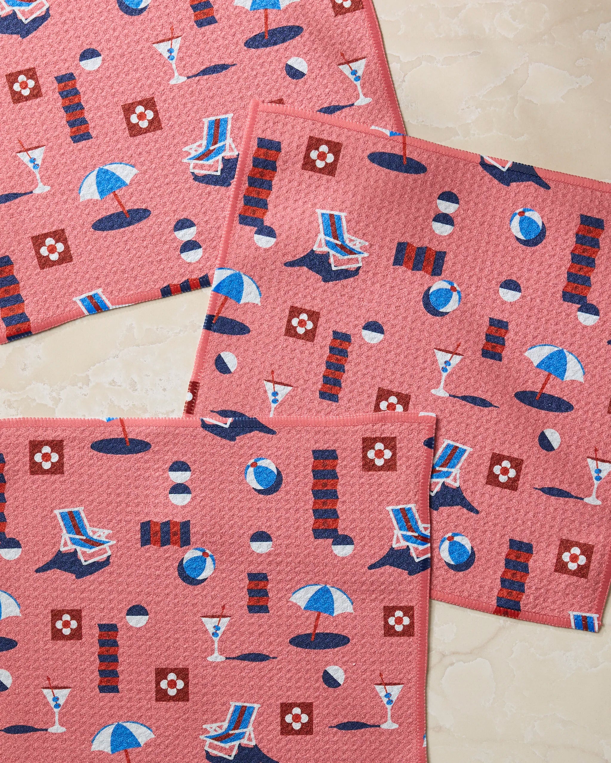 Three Geometry Geo Beach Day pink placemats, featuring a summer pattern of beach balls, umbrellas, lounge chairs, cocktails, and flowers, are displayed on a light marble surface—perfect alongside super-absorbent dishtowels.