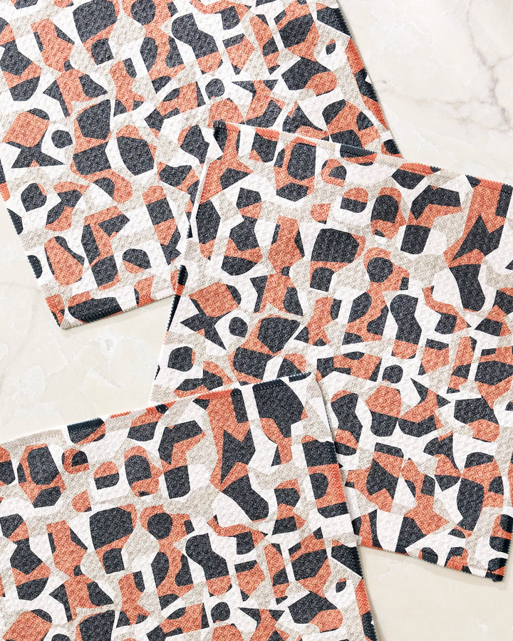 Four Geometric Leopard fabric swatches by Kris Ruff, made from recycled materials, show an abstract black, rust orange, and beige pattern. The overlapping swatches are displayed on a light marble surface.