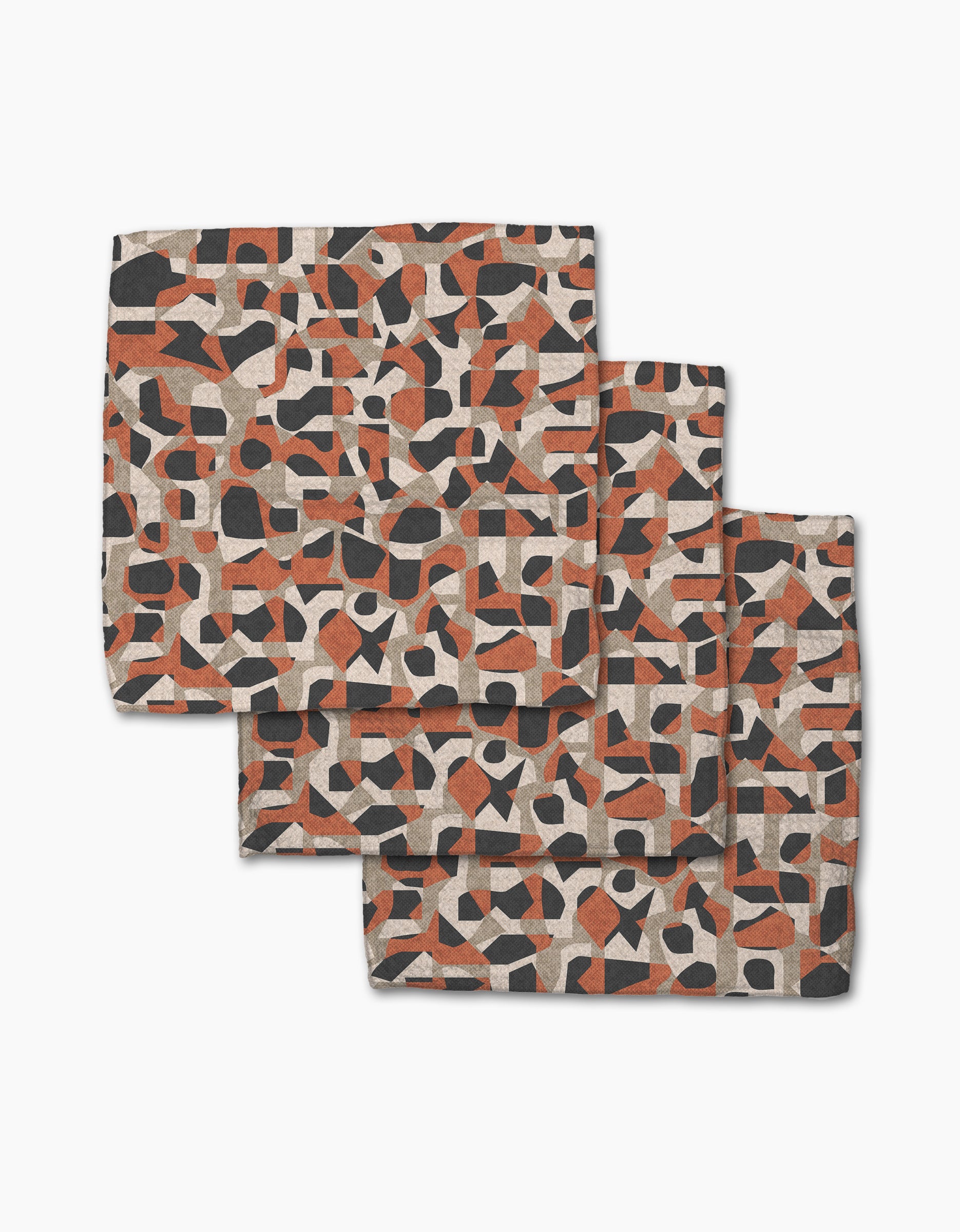 Three "Geometric Leopard" cloth napkins by Kris Ruff, featuring an abstract geometric pattern in black, rust orange, and beige, made from recycled materials and displayed in a staggered stack on a white background.