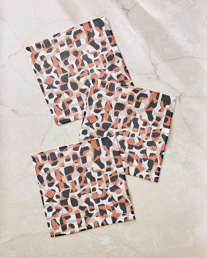 Three Geometric Leopard dishcloths by Kris Ruff, featuring a black, orange, and white abstract animal print, are arranged overlapping on a marble surface. This stylish set is made from recycled materials.