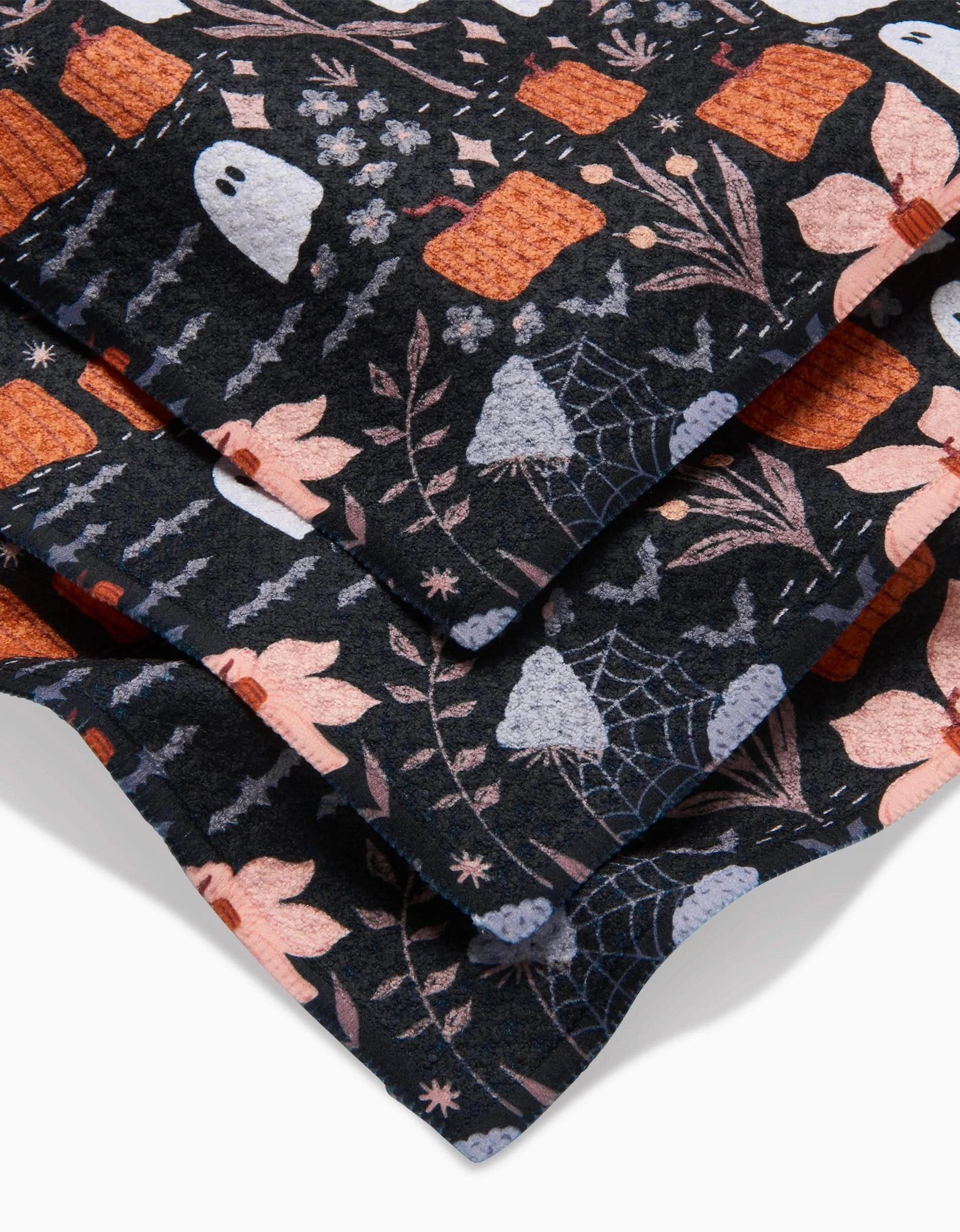 Close-up of Ghosts In The Midnight Garden by Charlie Rowan Designs—folded black fabric with white ghosts, cobwebs, orange books, and pink flowers. Perfect for super-absorbent, eco-friendly dish towels or dishcloths made from recycled materials.