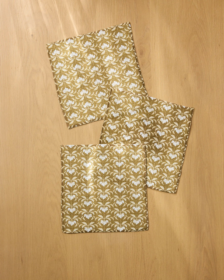 Three Gilded Bloom gold and white patterned absorbent dishcloths by Melissa Johnson are arranged on a light wood surface—two overlap at the corners, while the third is positioned separately above them.