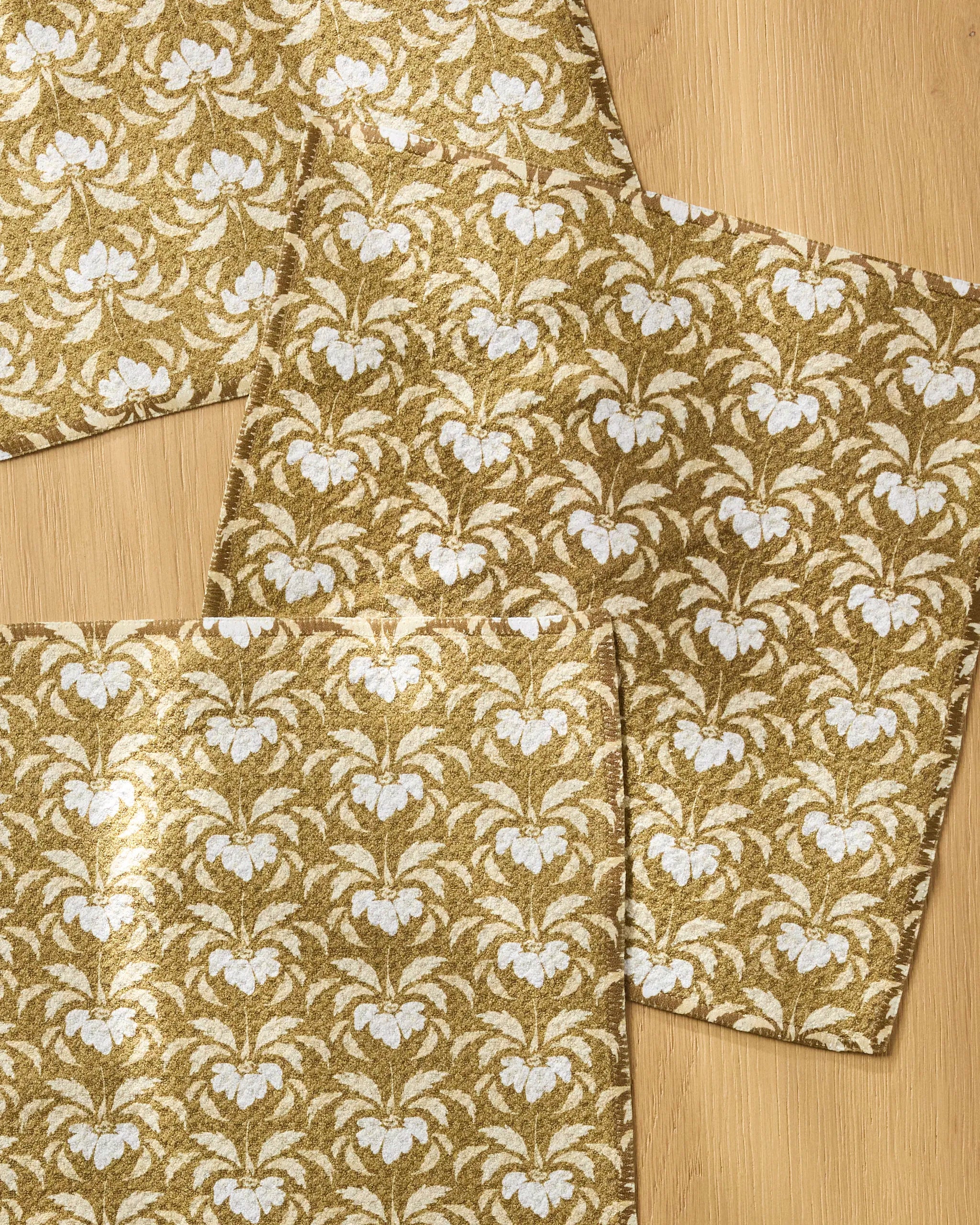Four Melissa Johnson Gilded Bloom gold placemats with a white floral pattern are arranged on a light wooden surface, some overlapping. Pair them with an absorbent dishcloth set for a stylish and practical table setting.
