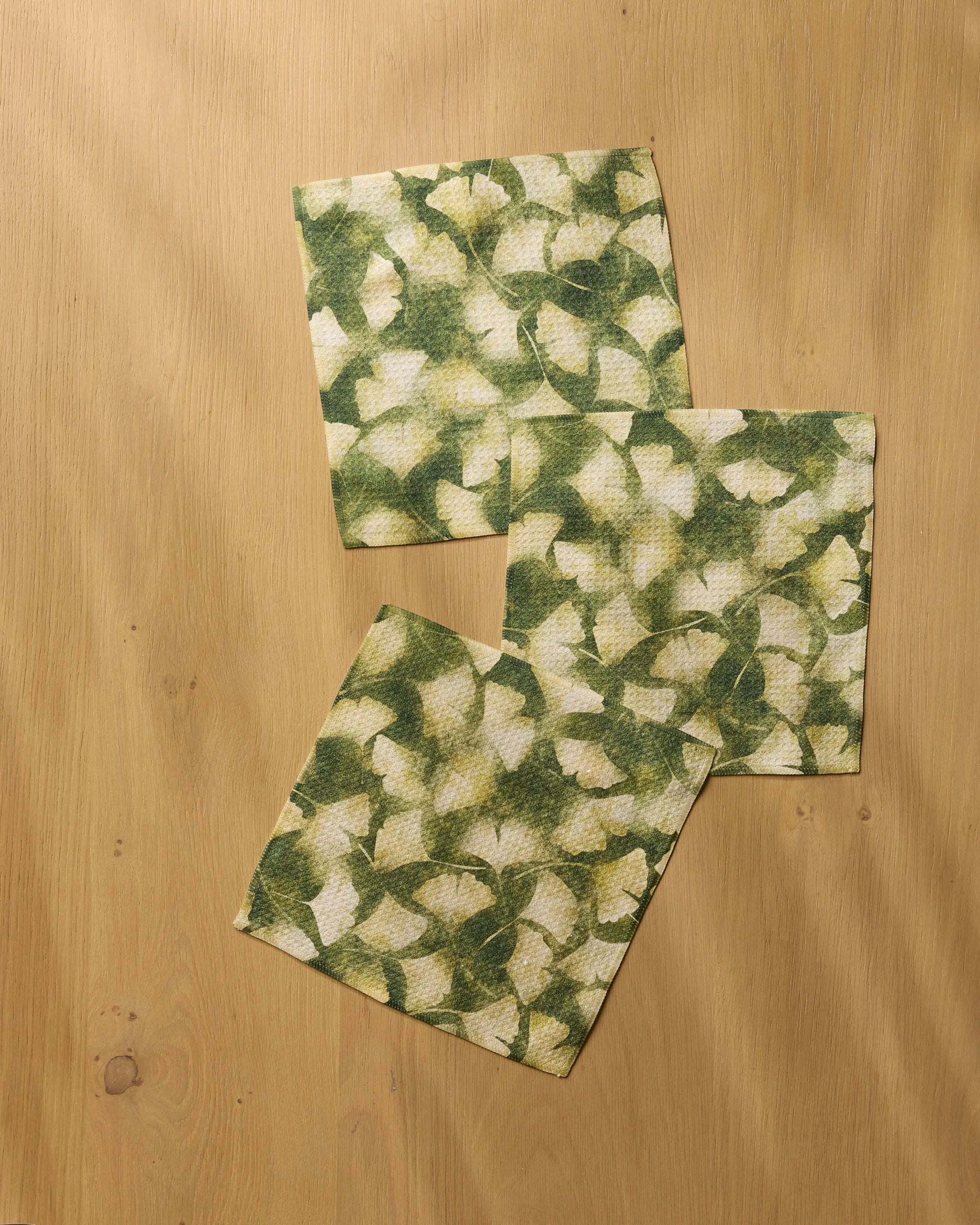 Three Gilded Ginkgo square napkins by Geometry, featuring a green and white leafy design and made from recycled materials, slightly overlap at the corners on a light wooden surface.