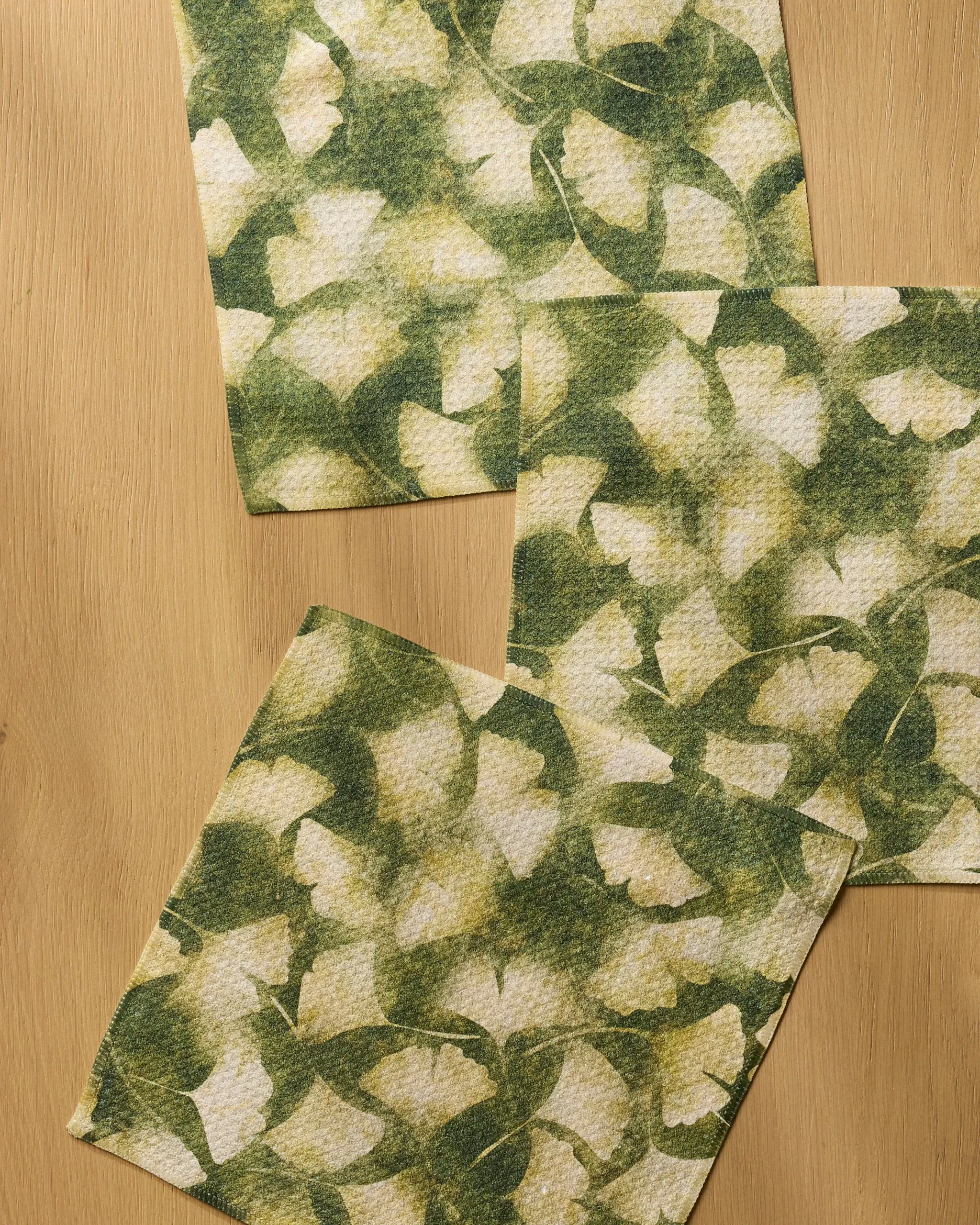 Three Geometry Gilded Ginkgo dishcloths, featuring a green and white leafy design and made from recycled materials, are laid out slightly overlapping on a light wooden surface.