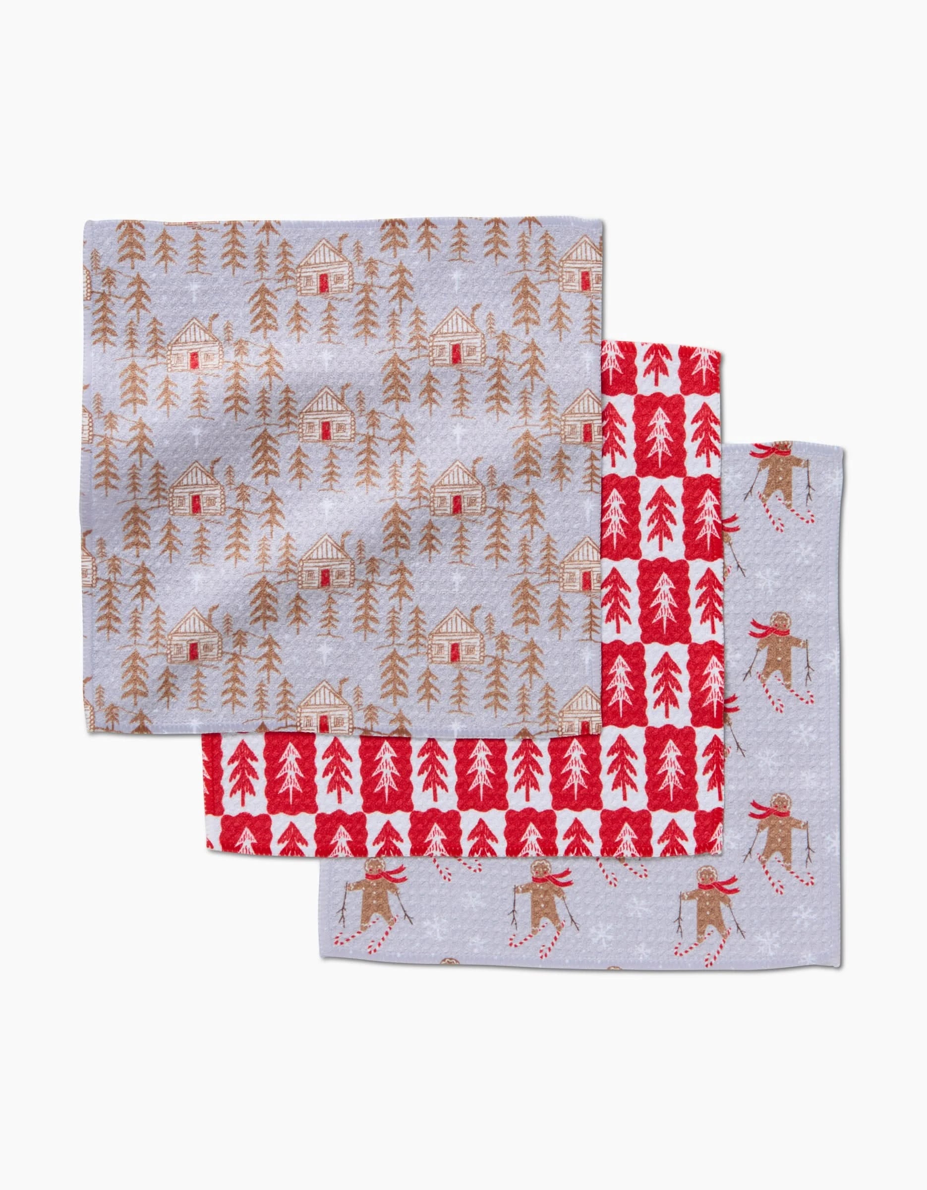 The Gingerbread Homestead set by Roxy Marj features three kitchen tea towels: brown cabins and trees, red trees on a white grid, and skiing dogs in red scarves—all on light grey Geoweave fabric with winter-themed prints.