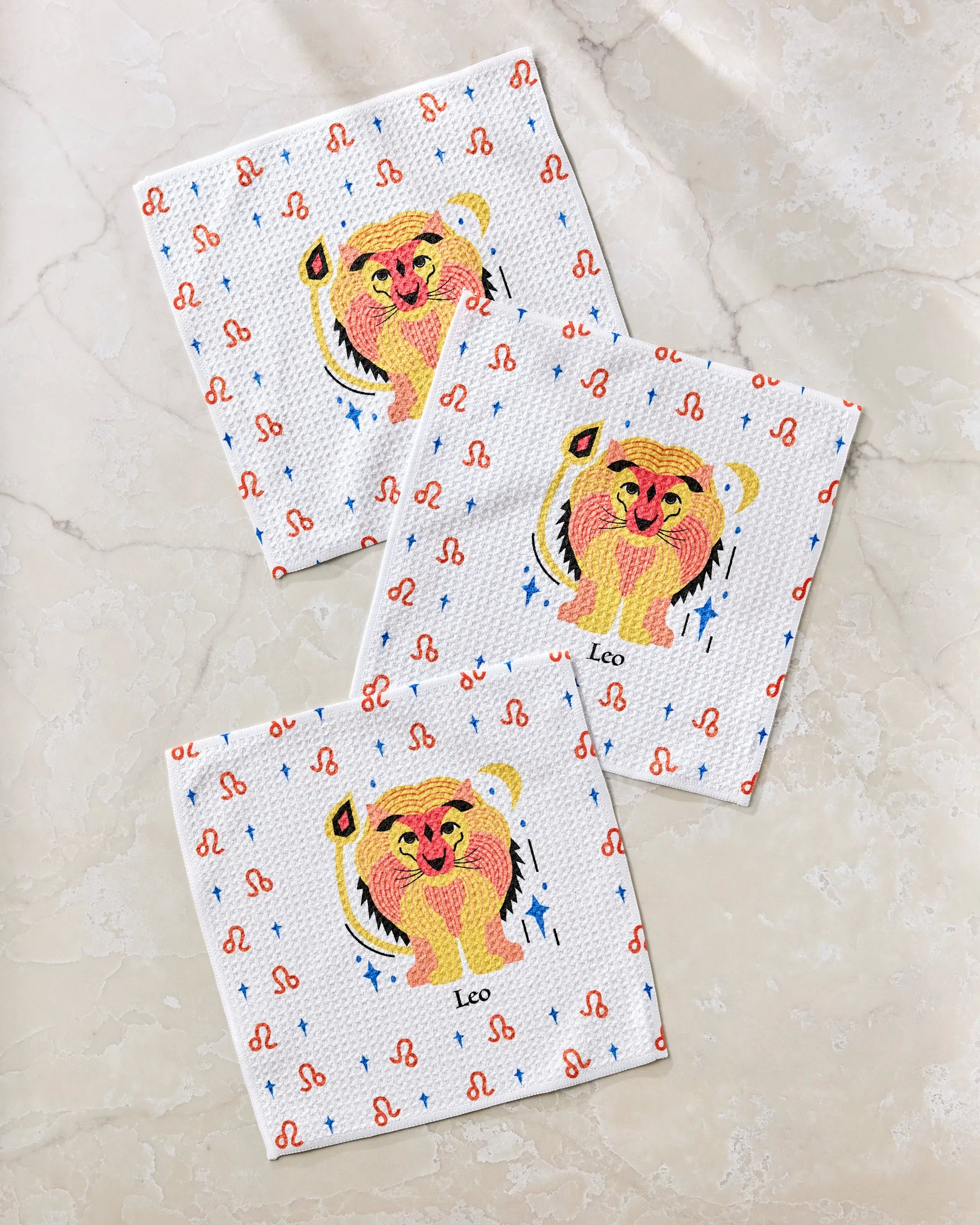 Three white Glowing Leo dishcloths by Heather Zhou feature a playful lion, the word "Leo," red zodiac symbols, and blue stars. These reusable, eco-friendly towels are arranged on a light marble surface.